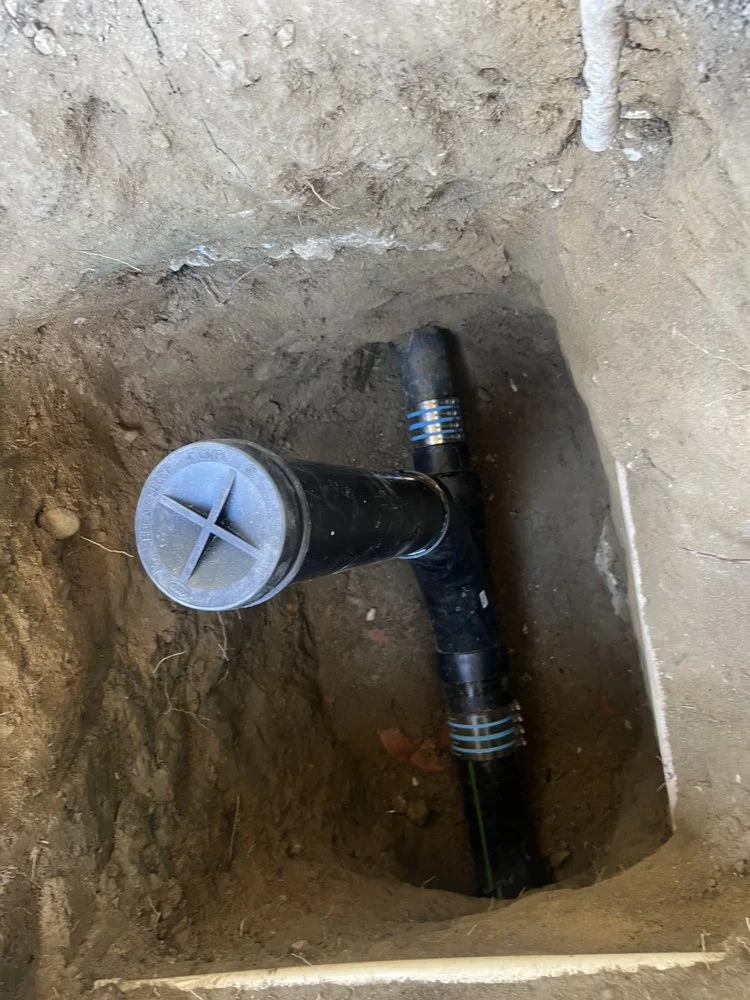 Close-up of a plumbing pipe inside a dug hole in the ground, with a black pipe and a gray plastic cap on top, surrounded by dirt.