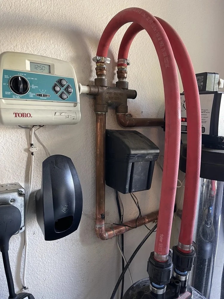 A wall-mounted water heater timer, plumbing pipes, and a black water filter system with pink flexible hoses connected to it.