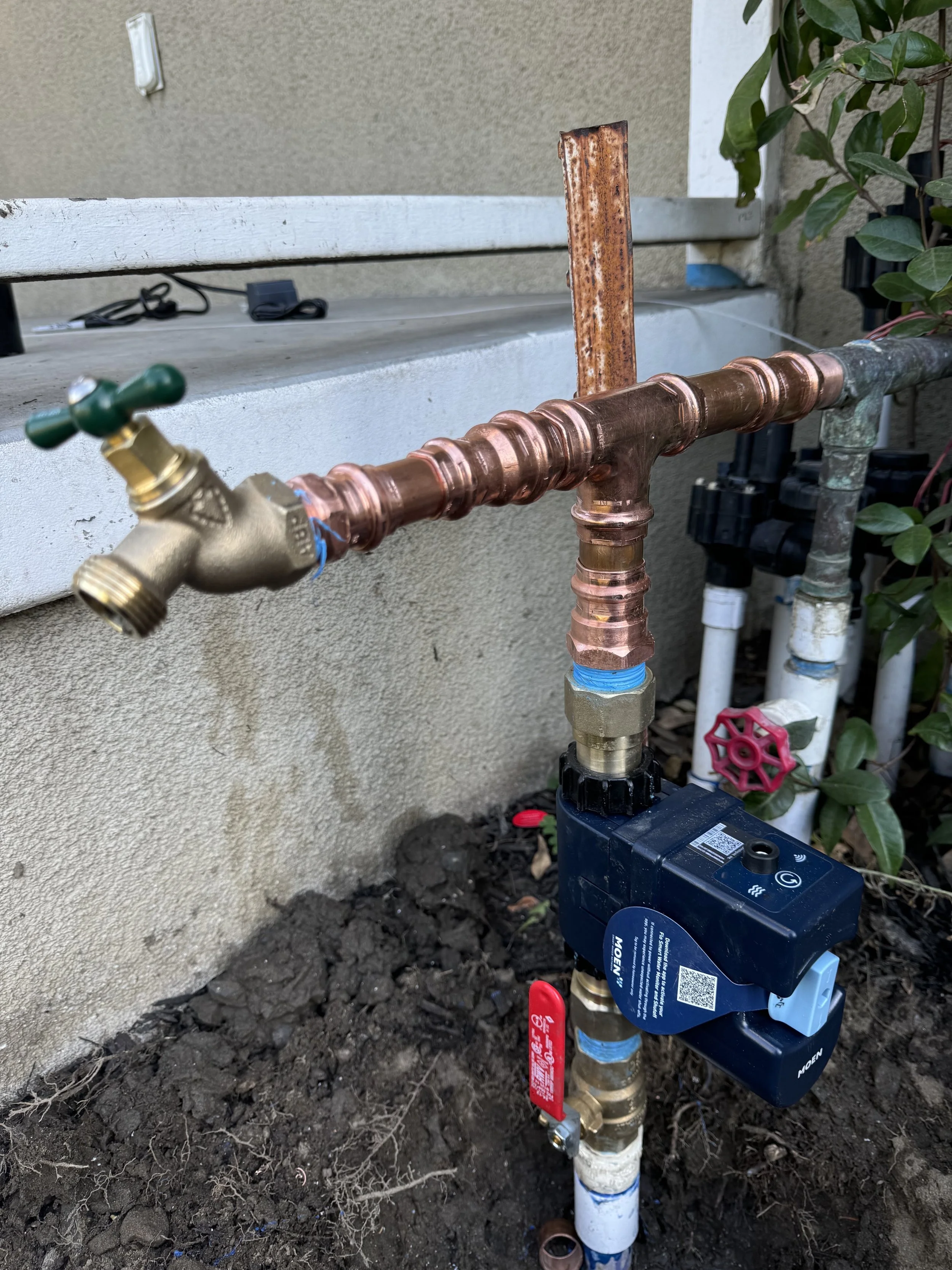 outdoor plumbing system with copper pipes, a brass valve, and a black electronic valve controller, set against a concrete wall and soil ground, with plants nearby.