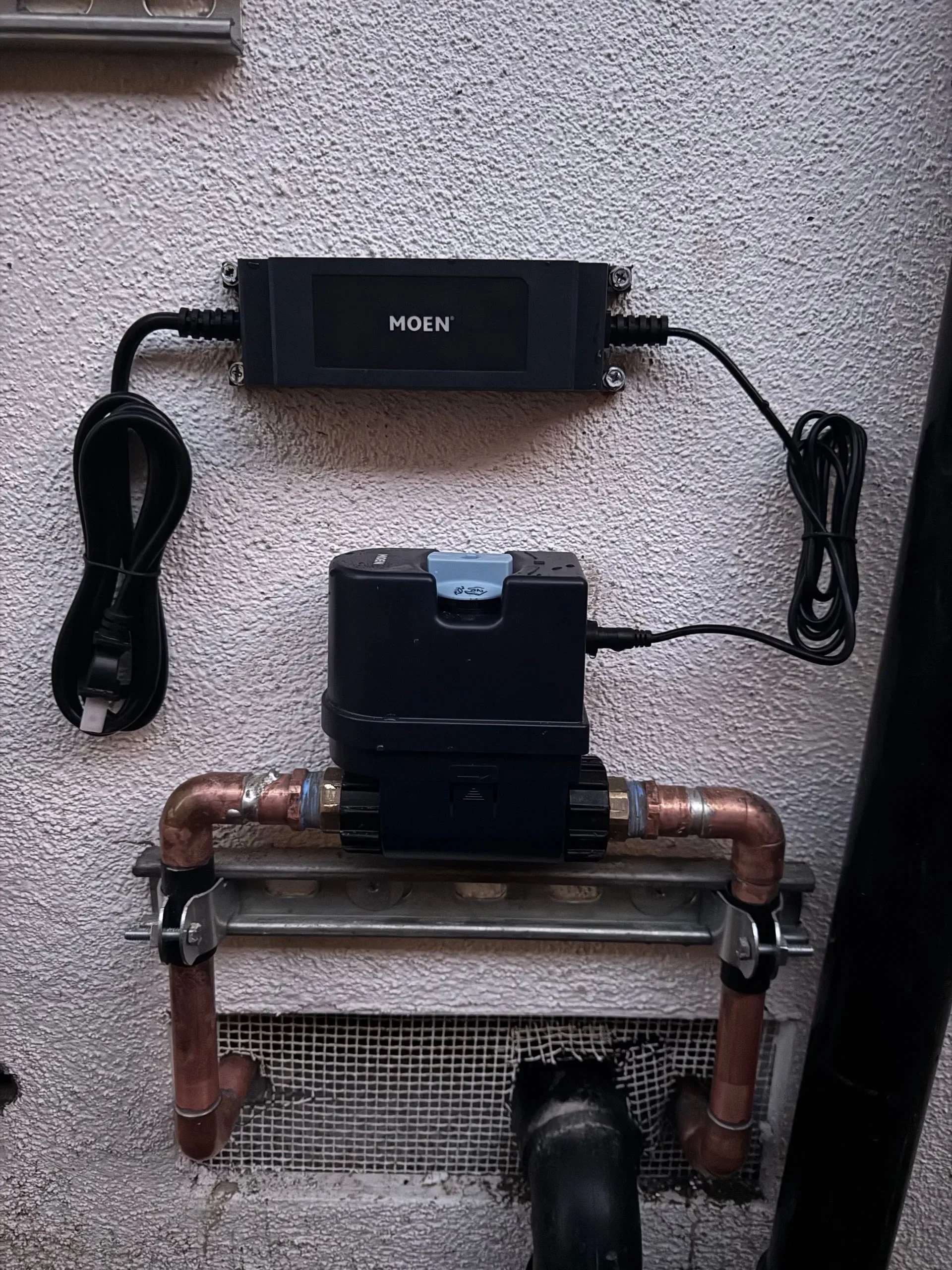 A plumbing system installed on an exterior wall, comprising a Moen control box, a black pump, copper pipes, and a vent opening.