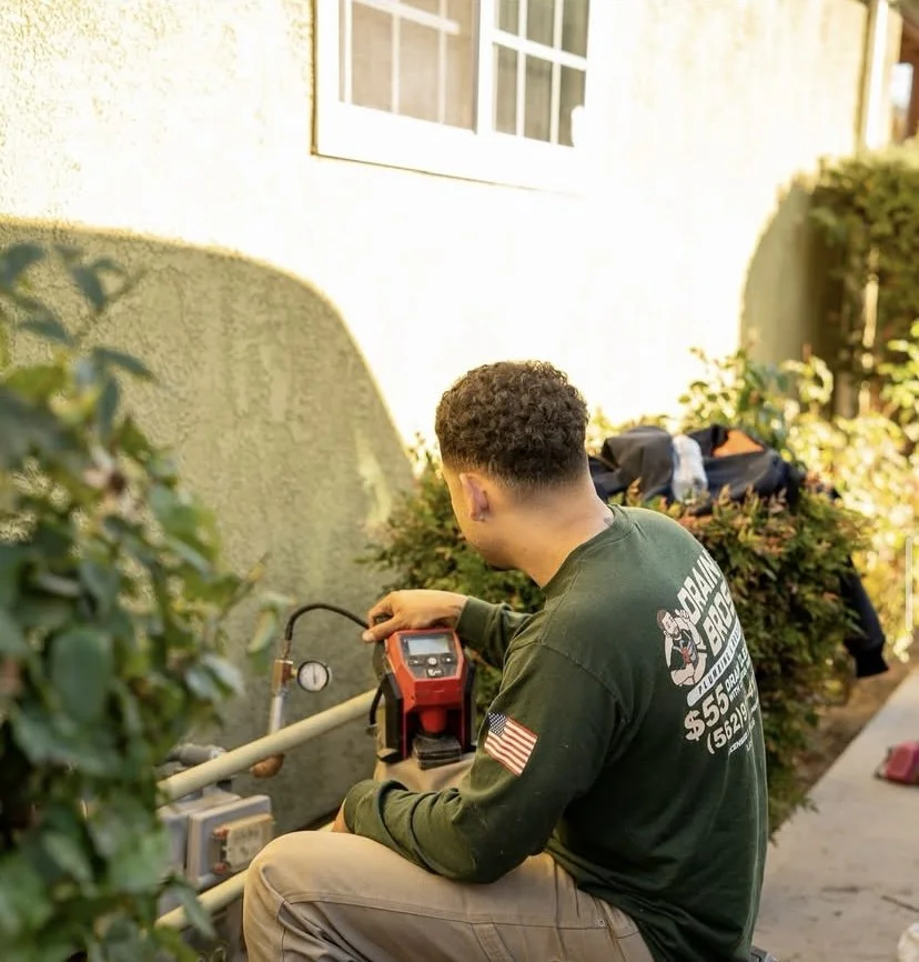 A worker kneeling next to a wall with a pressure test gauge, holding a device, with greenery and a window in the background.