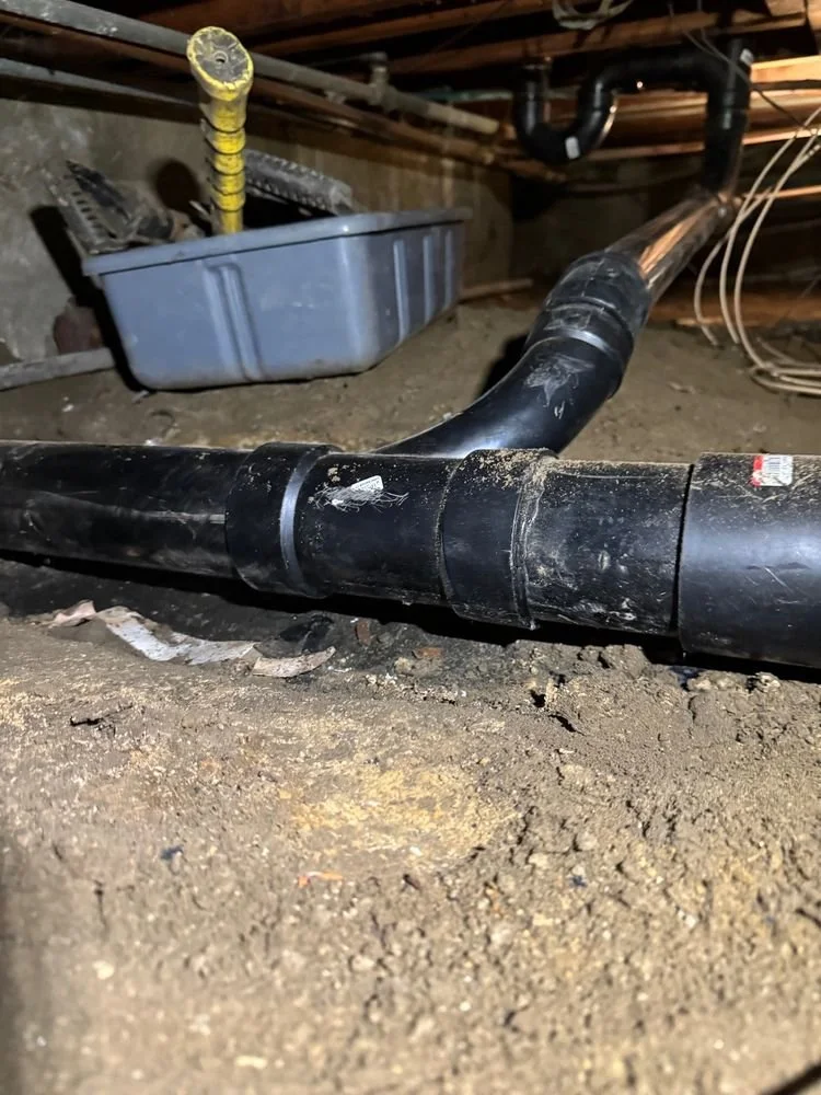 Black plastic perforated pipe on dirt floor in a crawl space with a gray plastic container and a yellow-handled tool in the background.