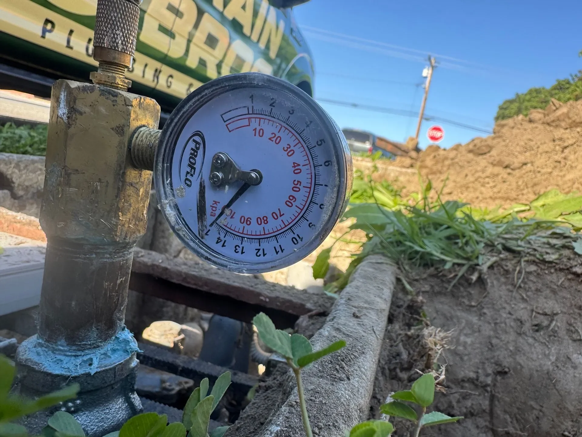 A close-up of a pressure gauge in a garden, with a background of soil, green plants, a blue sky, and a sign that reads 'PLUM'.