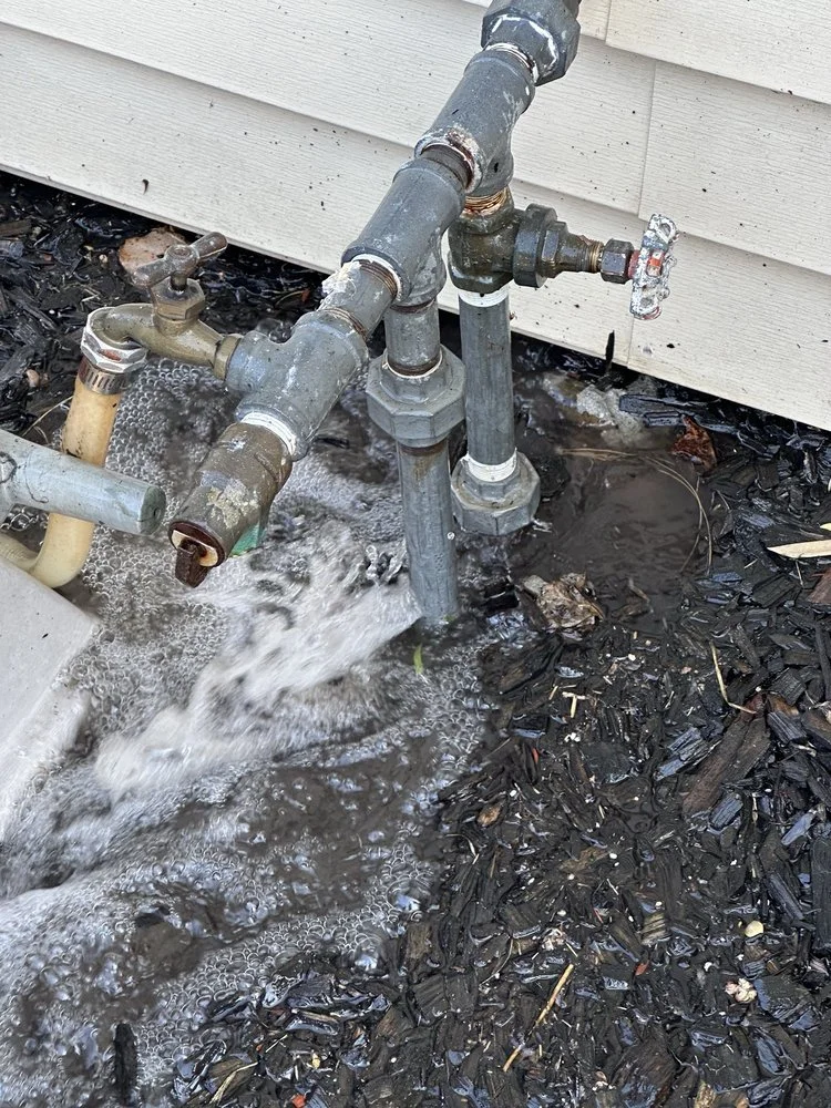 Outdoor water pipe with water flowing out onto mulch and soil, next to the house siding.