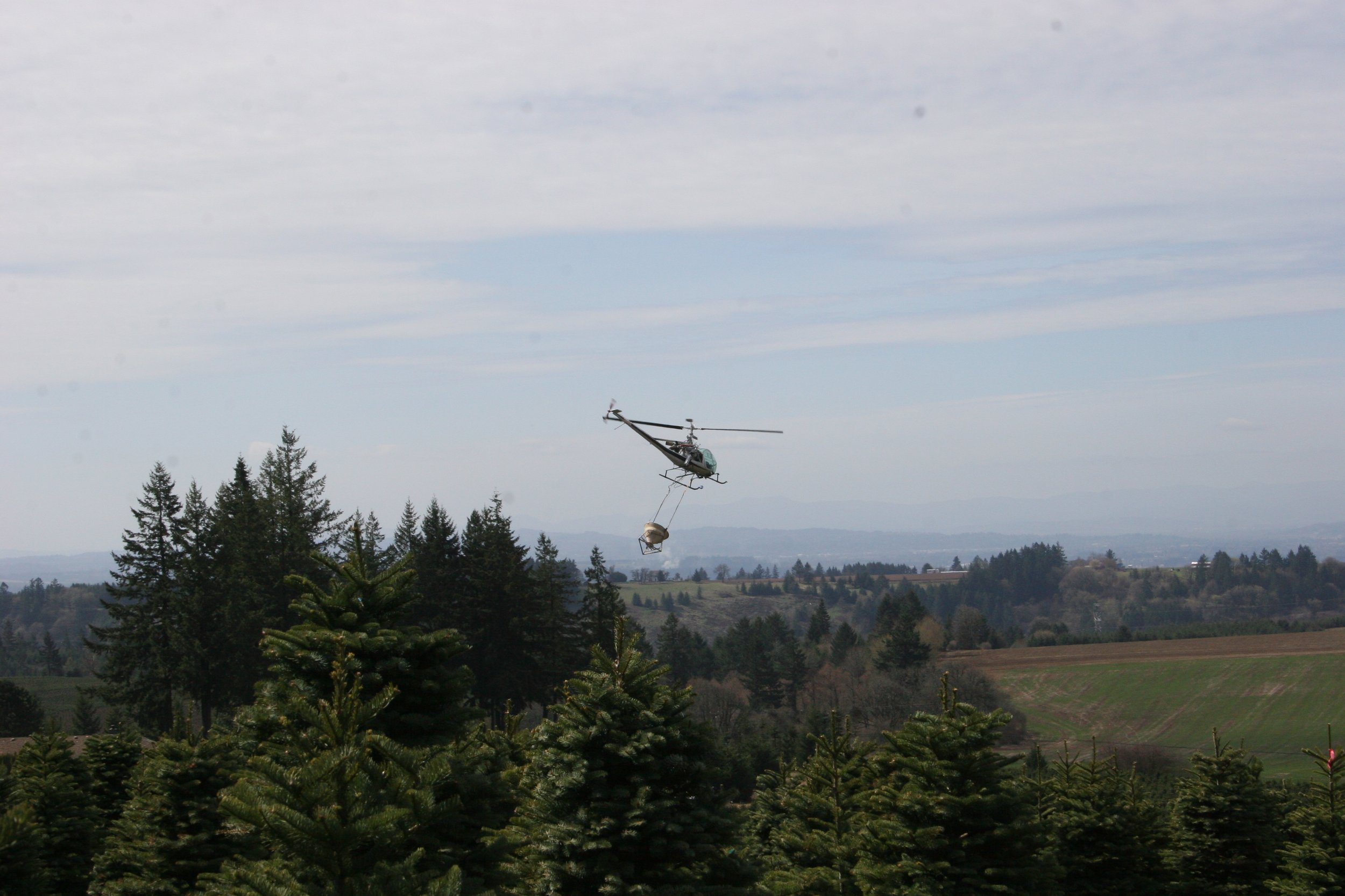 A helicopter flying over a forest carries a bucket hanging from a cable, with a cloudy sky above and a landscape of trees and distant hills below.