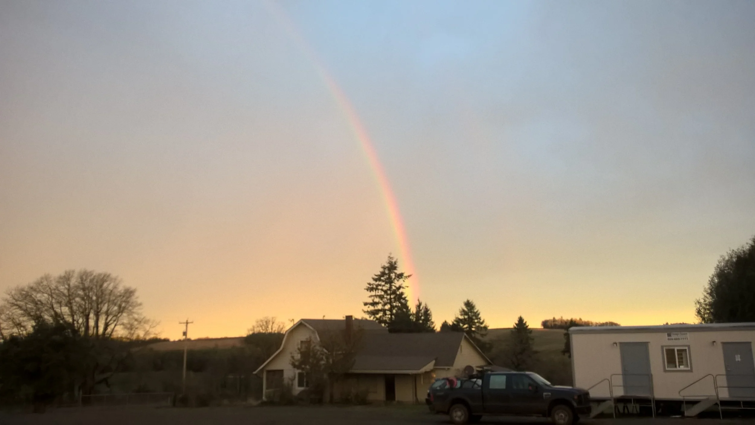 A rainbow arcing in the sky above a rural landscape during sunset, with houses, trees, and a parked vehicle in the foreground.
