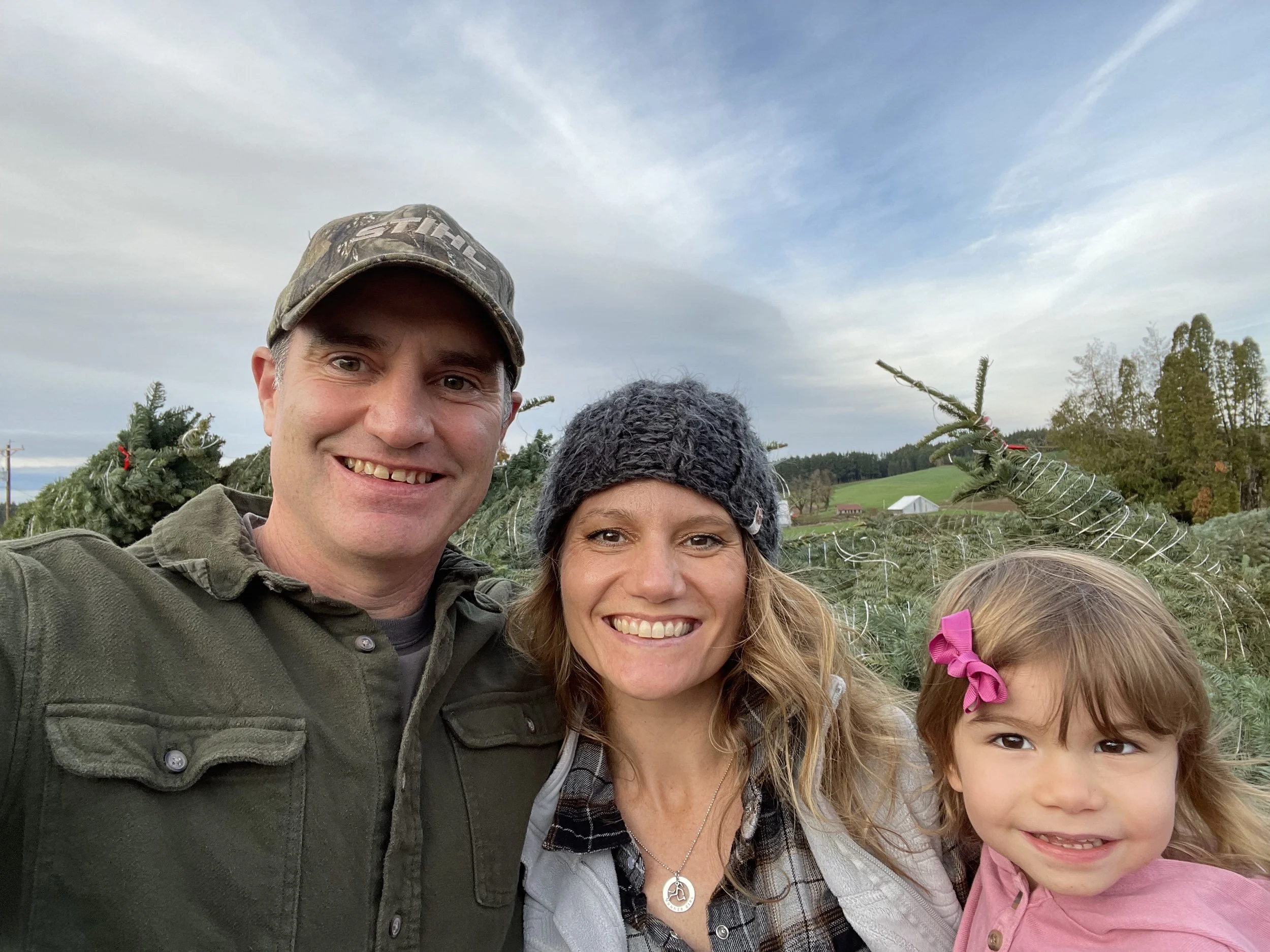 A family of three taking a selfie outdoors in front of a Christmas tree farm, with trees and green hills in the background, on a partly cloudy day.