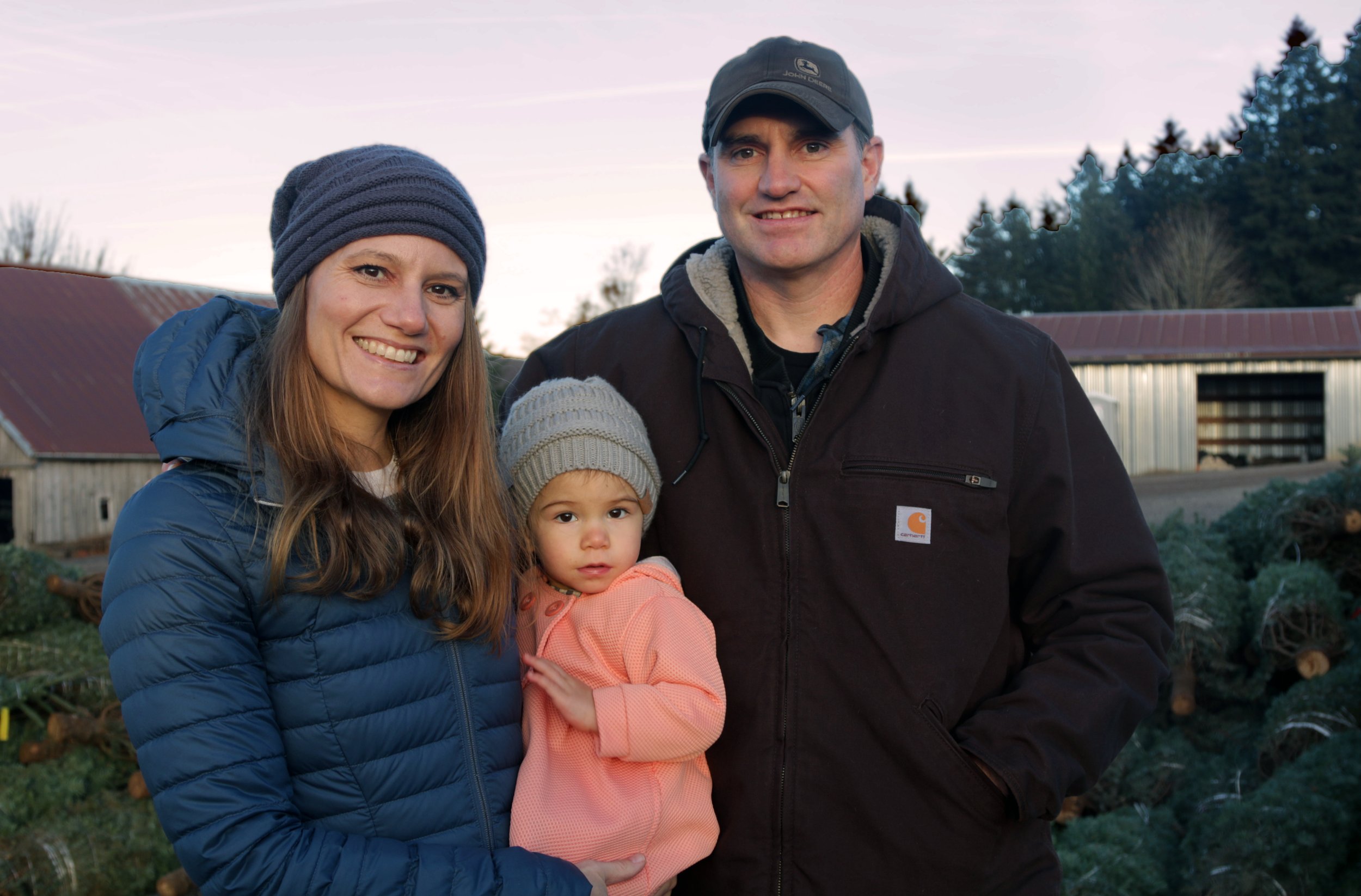 A smiling family of three stands outdoors at a farm with trees and barn in the background, during sunset. The woman is wearing a blue jacket and gray knit hat. The man is wearing a black jacket and baseball cap. The little girl, in an orange jacket a