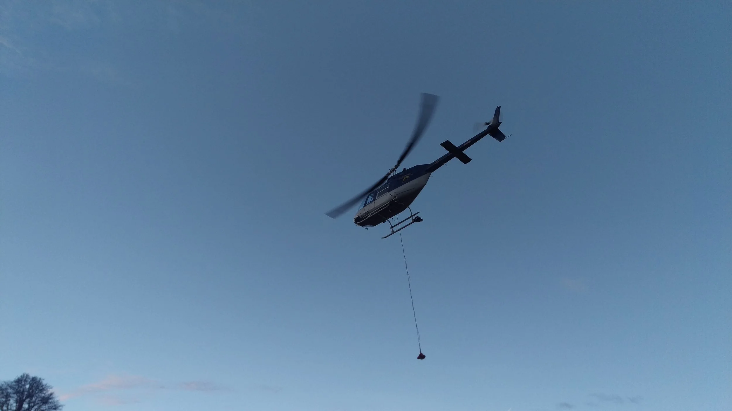 A helicopter flying in the sky with a bucket hanging from a cable underneath it.