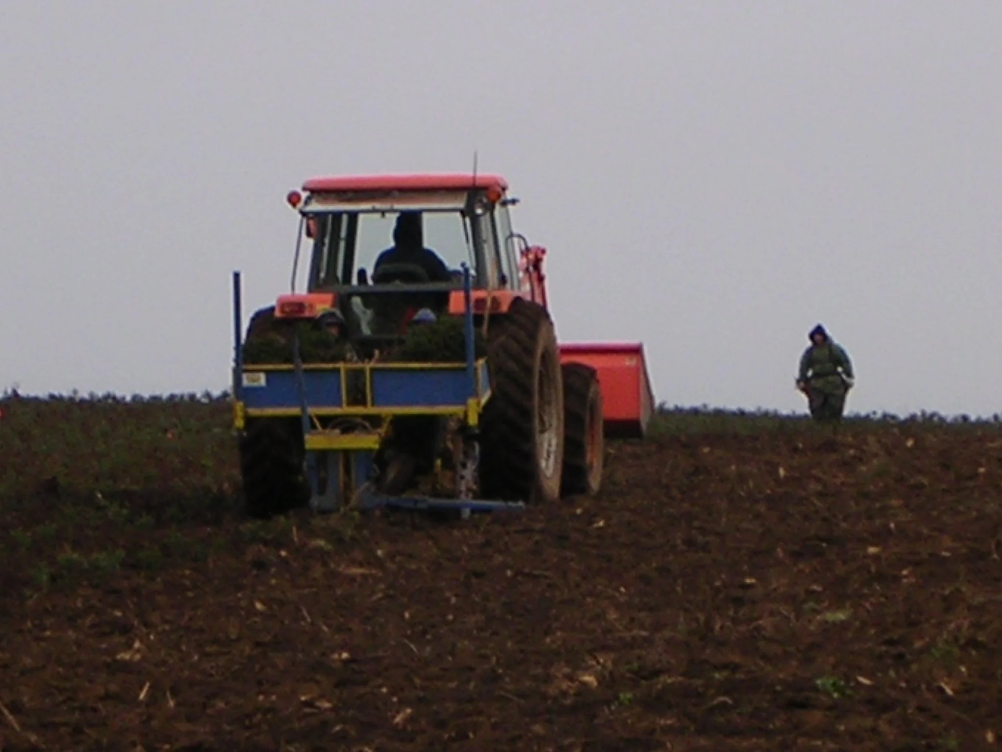 A tractor on a tilled farm field with a person walking in the background