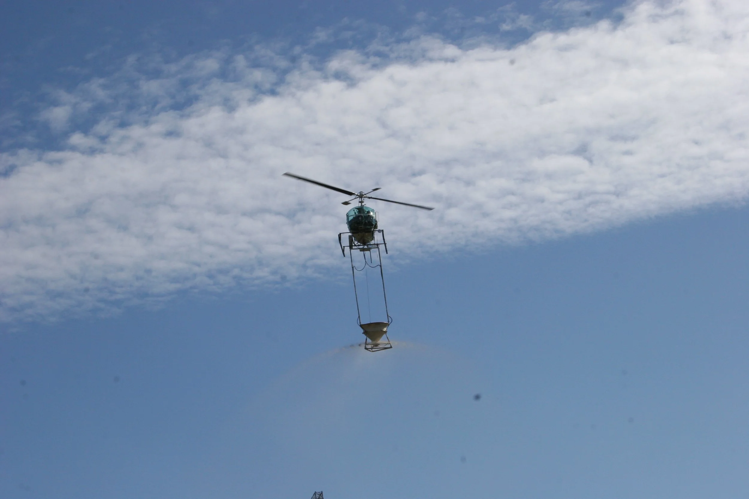 A helicopter carrying a bucket, flying in a partly cloudy sky.