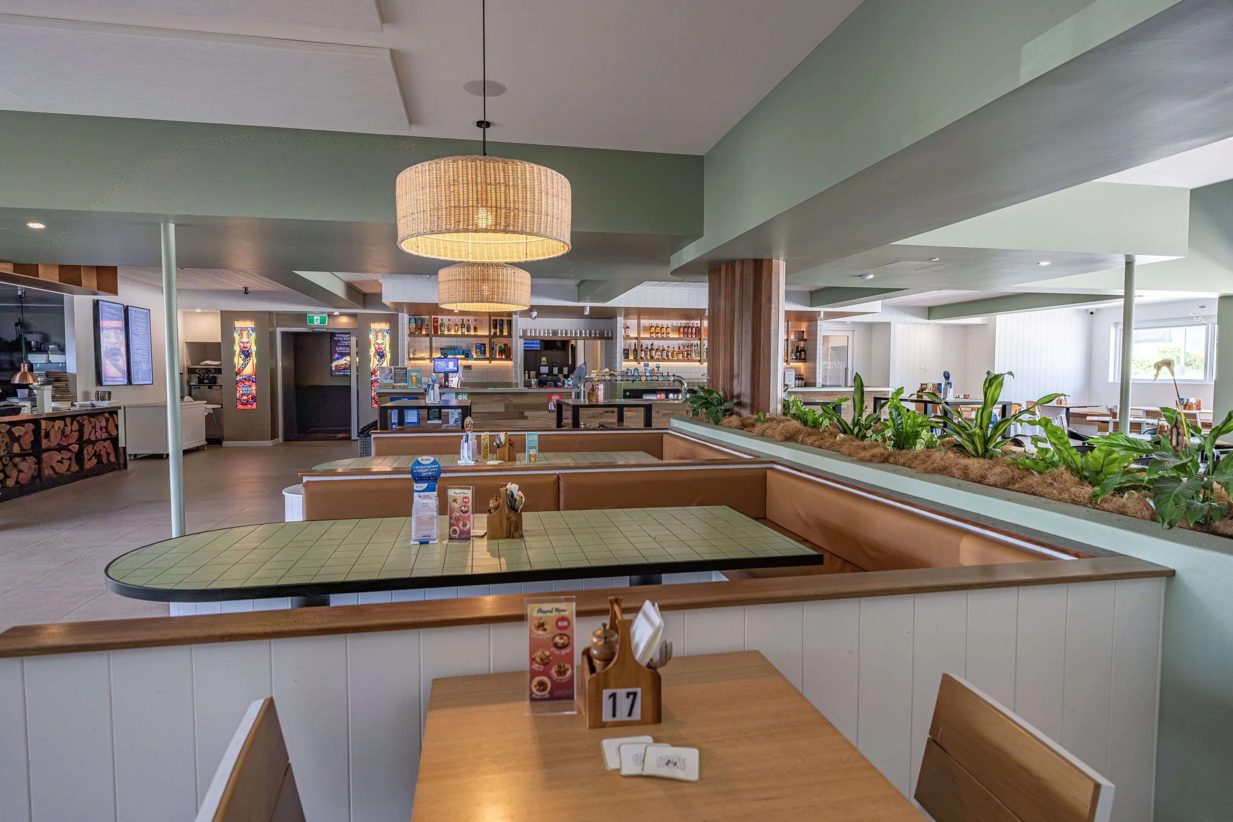 Modern bistro with green and white walls, wooden accents, tropical plants, and brown booths, illuminated by hanging woven light fixtures.