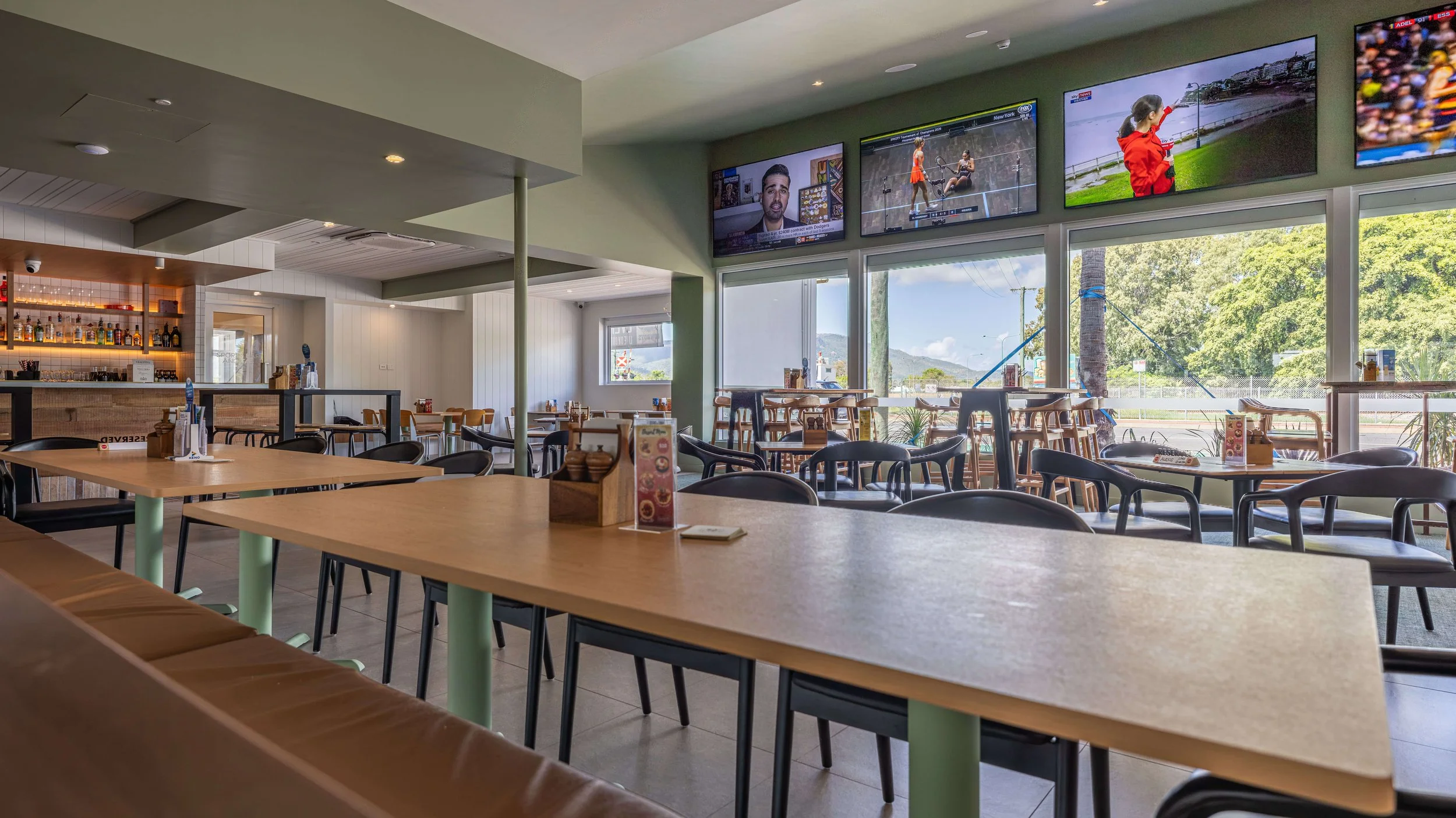Modern bistro interior with tables, chairs, a bar, and multiple wall-mounted television screens showing sports and news, with large windows letting in natural light and providing a view of trees and outdoor scenery.