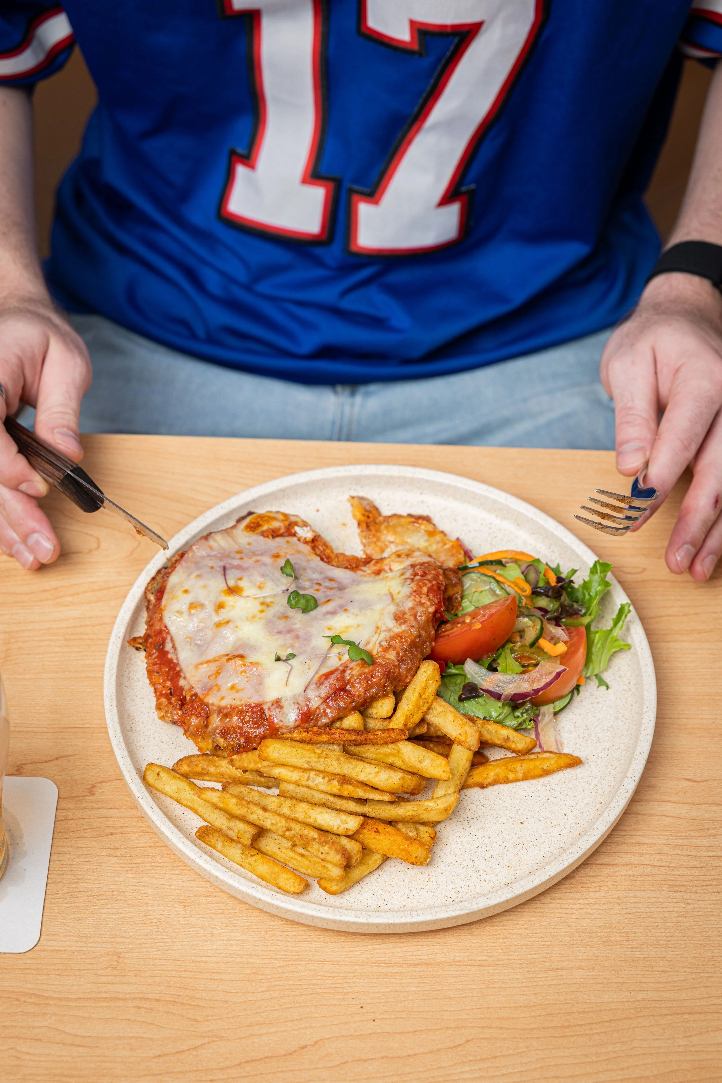 A person sits at a wooden table with a plate parmigiana, fries and salad.