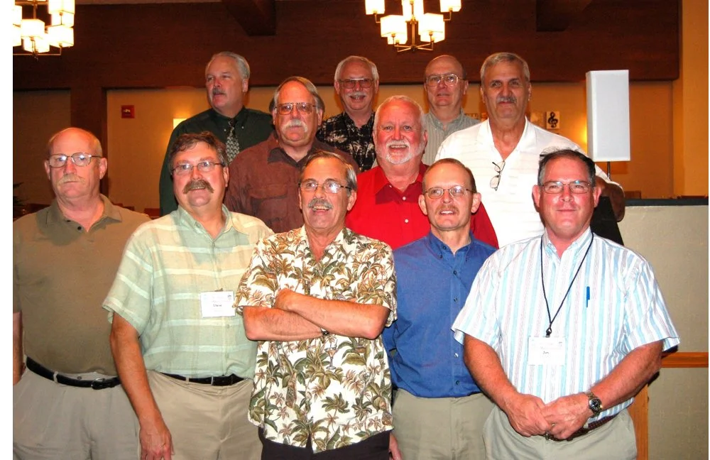 Group of eleven men gathered in a room with wooden walls and warm lighting, some wearing name tags, posing for a photo.