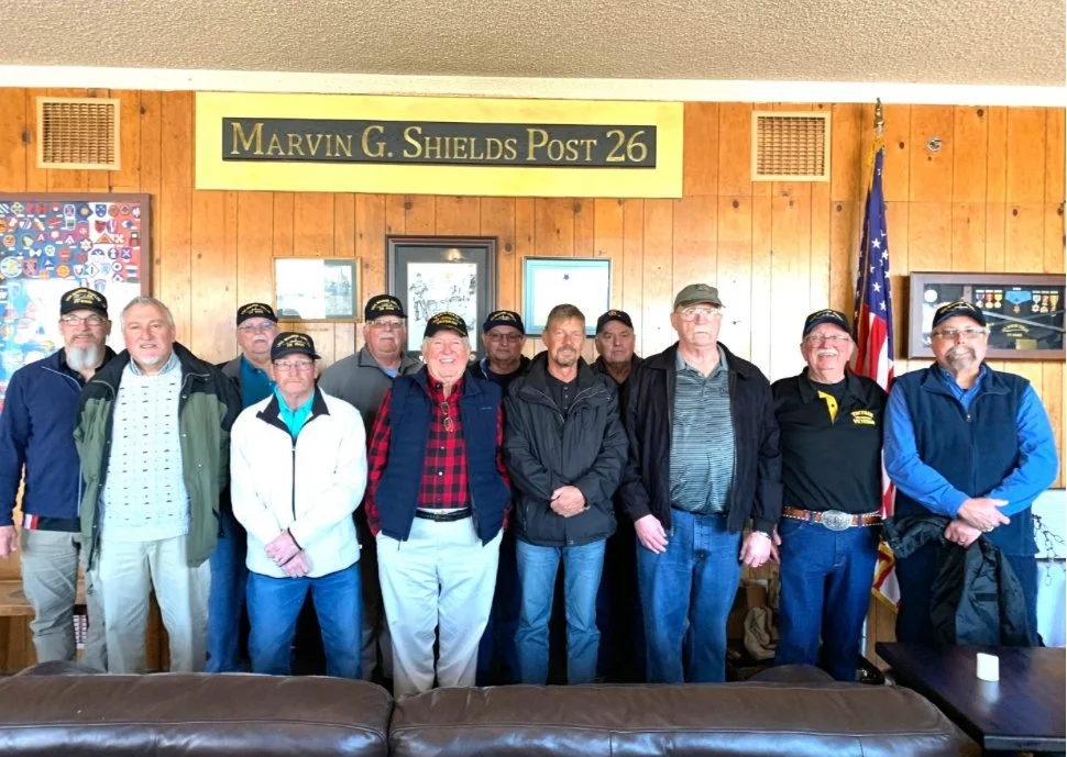 Group of nine men standing inside a wood-paneled room, with a sign that reads 'Marvin G. Shields Post 26' and an American flag in the background.