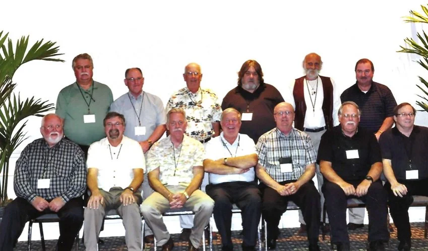 Group of 11 men posing for a photo, seated and standing, in a room with white background and tall potted plants.