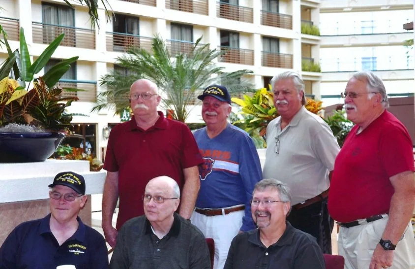 Seven older men gathered at an indoor garden or atrium with tropical plants and a multi-story building visible in the background. Four men are standing, three are sitting, and they are dressed casually, with some wearing hats. They appear to be enjoying a social event or reunion.