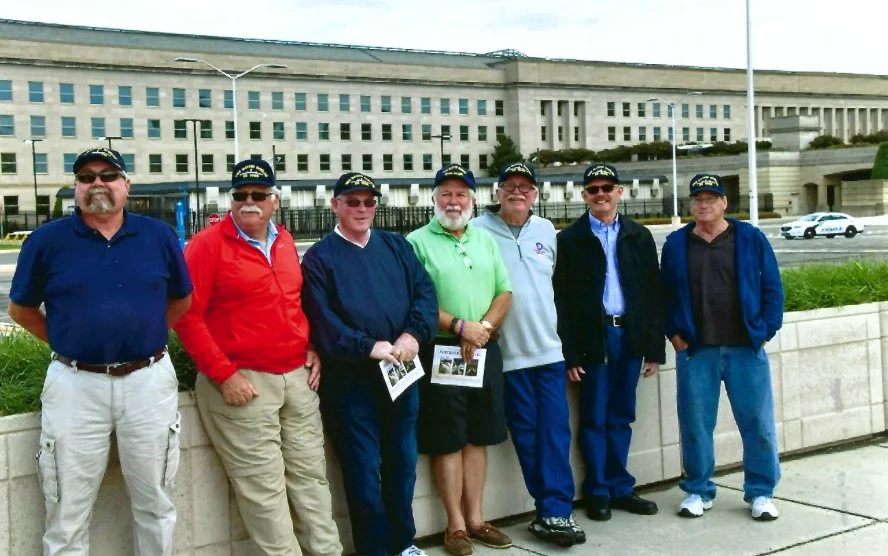 Seven men standing outdoors in front of a large government building, wearing casual clothes and Navy veteran caps, some holding pamphlets.