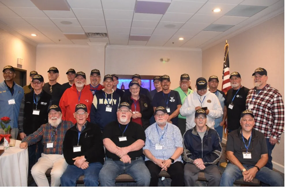Group of men and women wearing matching black baseball caps, some with navy or black jackets, gathered indoors for a group photo near an American flag.