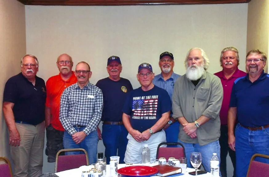 A group of ten men standing in a line inside a room, dressed casually, some smiling, with a table in front of them holding food and drinks.