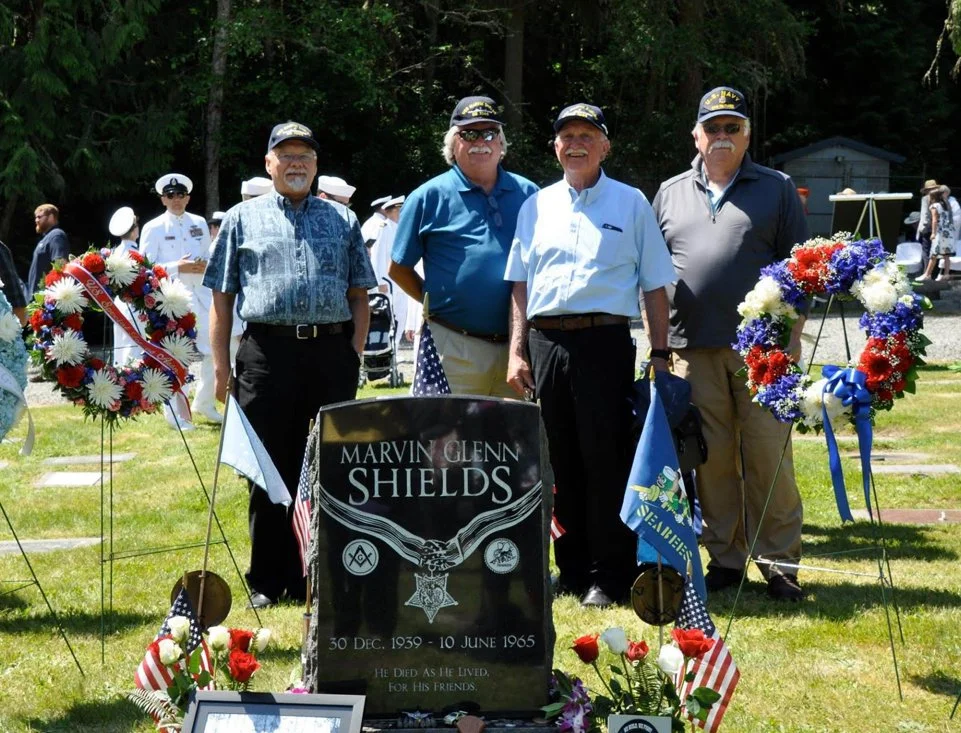 Four men standing at a gravesite during a memorial event for Marvin Glenn Shields, who served from December 30, 1939, to June 10, 1965. The grave is decorated with American flags and floral wreaths, and some individuals are holding small flags. Background shows more people, some in military uniforms, and trees.