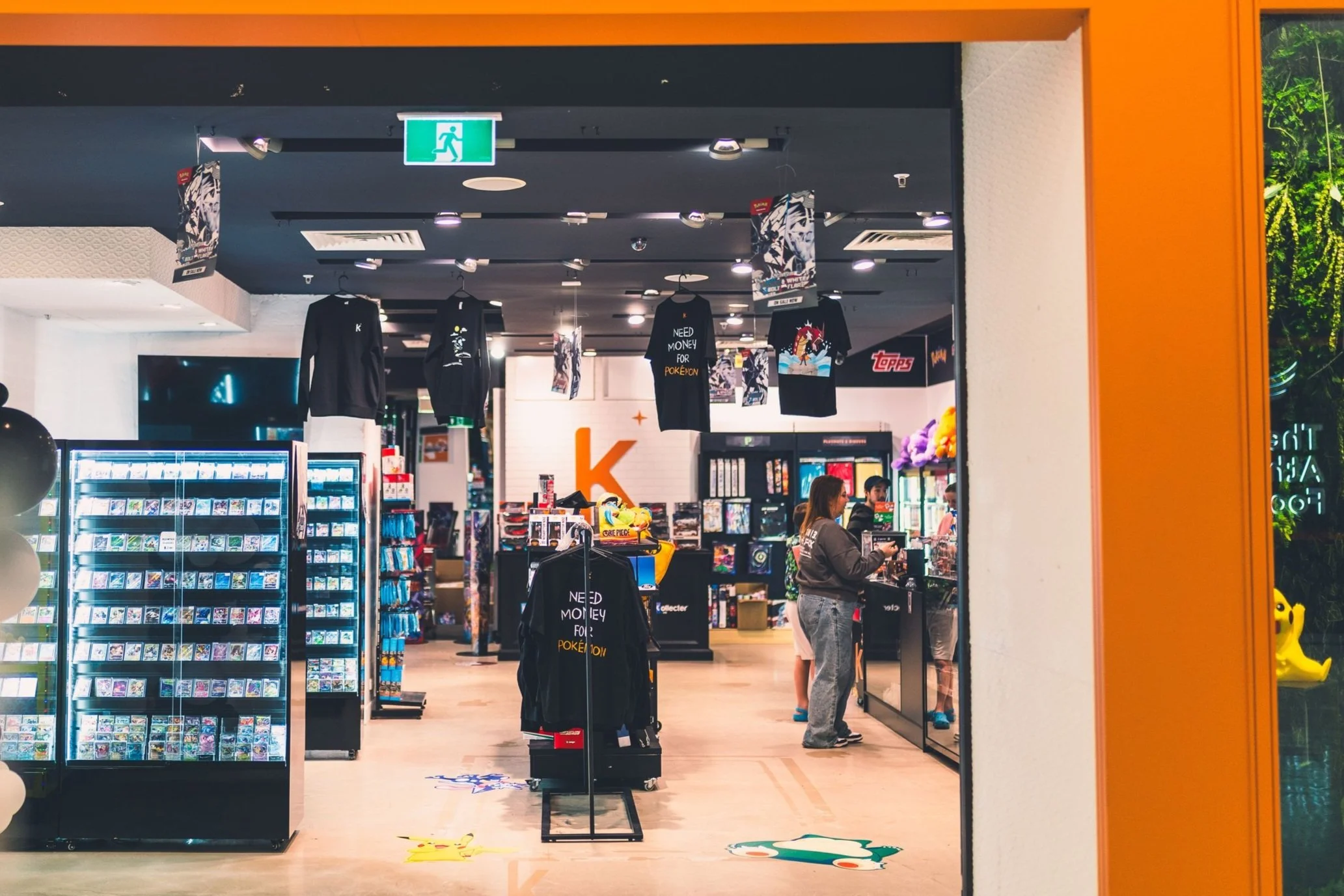 Inside a retail store with shelves of merchandise, clothing hanging from the ceiling, and customers at the checkout counter, illuminated by bright lights.