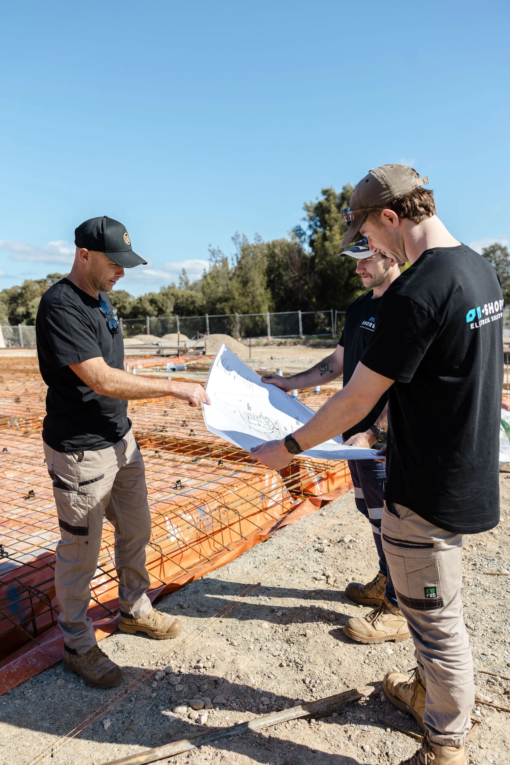 Construction workers review blueprints at a building site with exposed rebar foundation.