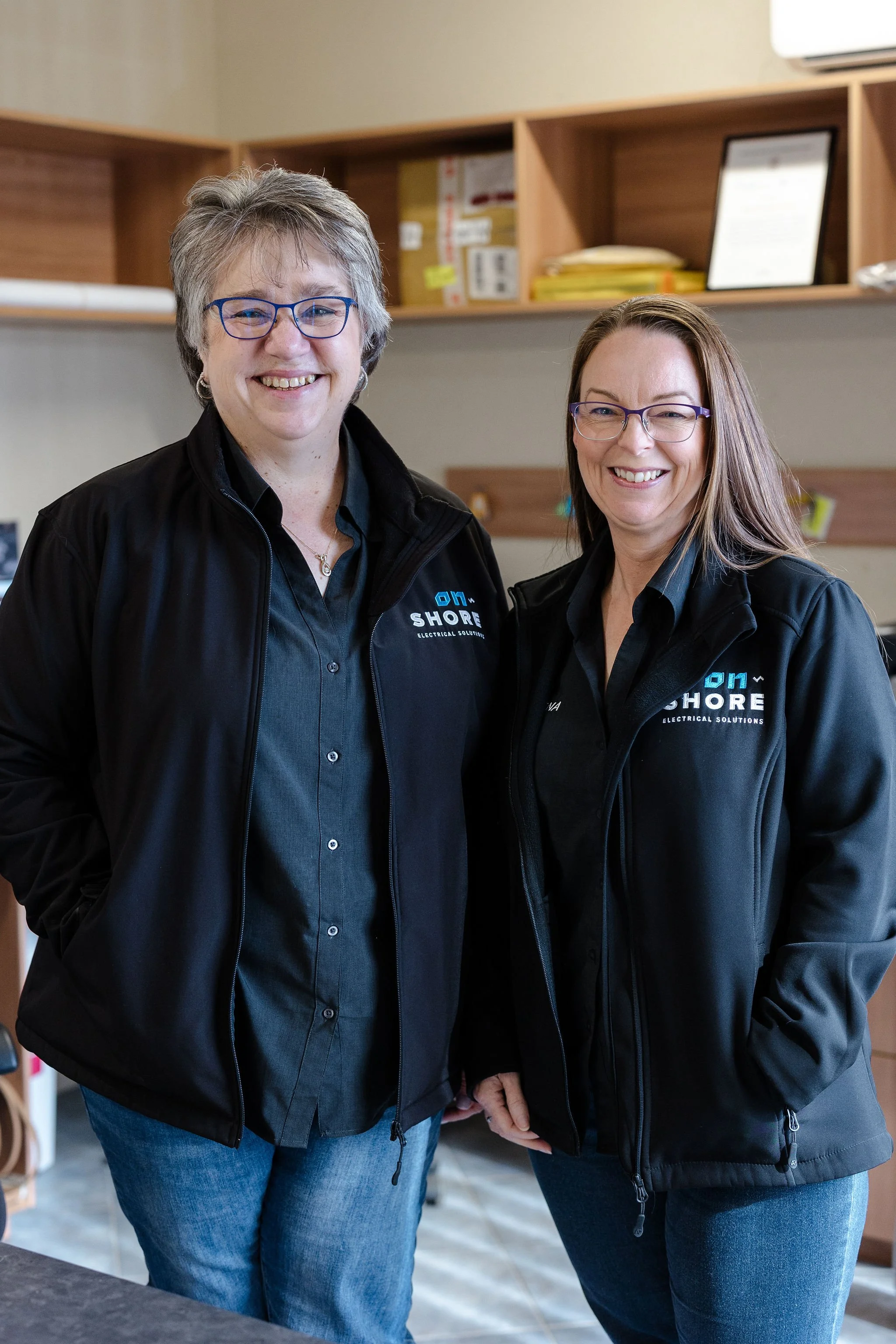 Two smiling women in black Shore Electrical Solutions jackets pose together indoors.