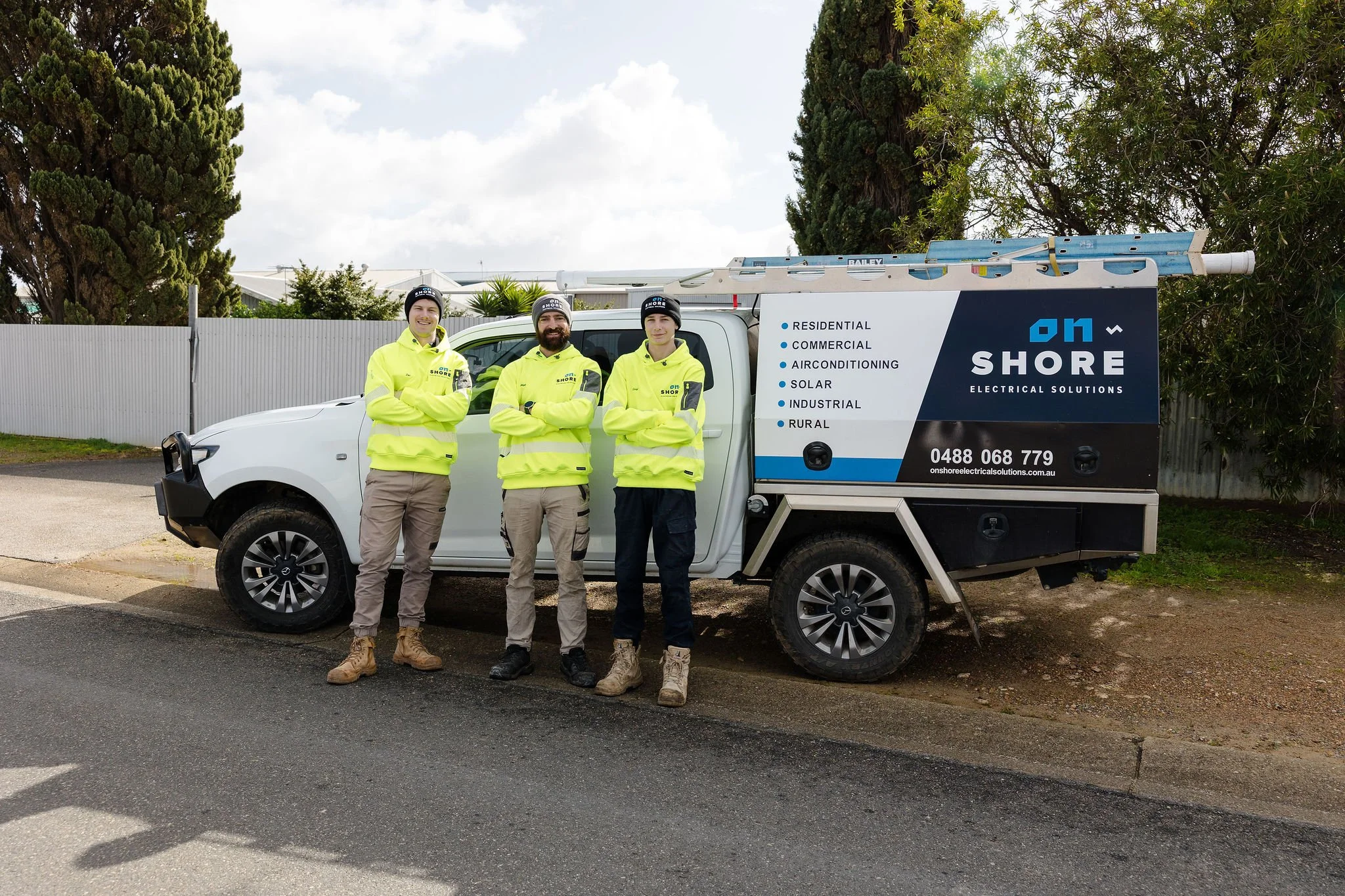 Three electricians in high-visibility uniforms stand beside an On Shore Electrical Solutions service truck with ladders.