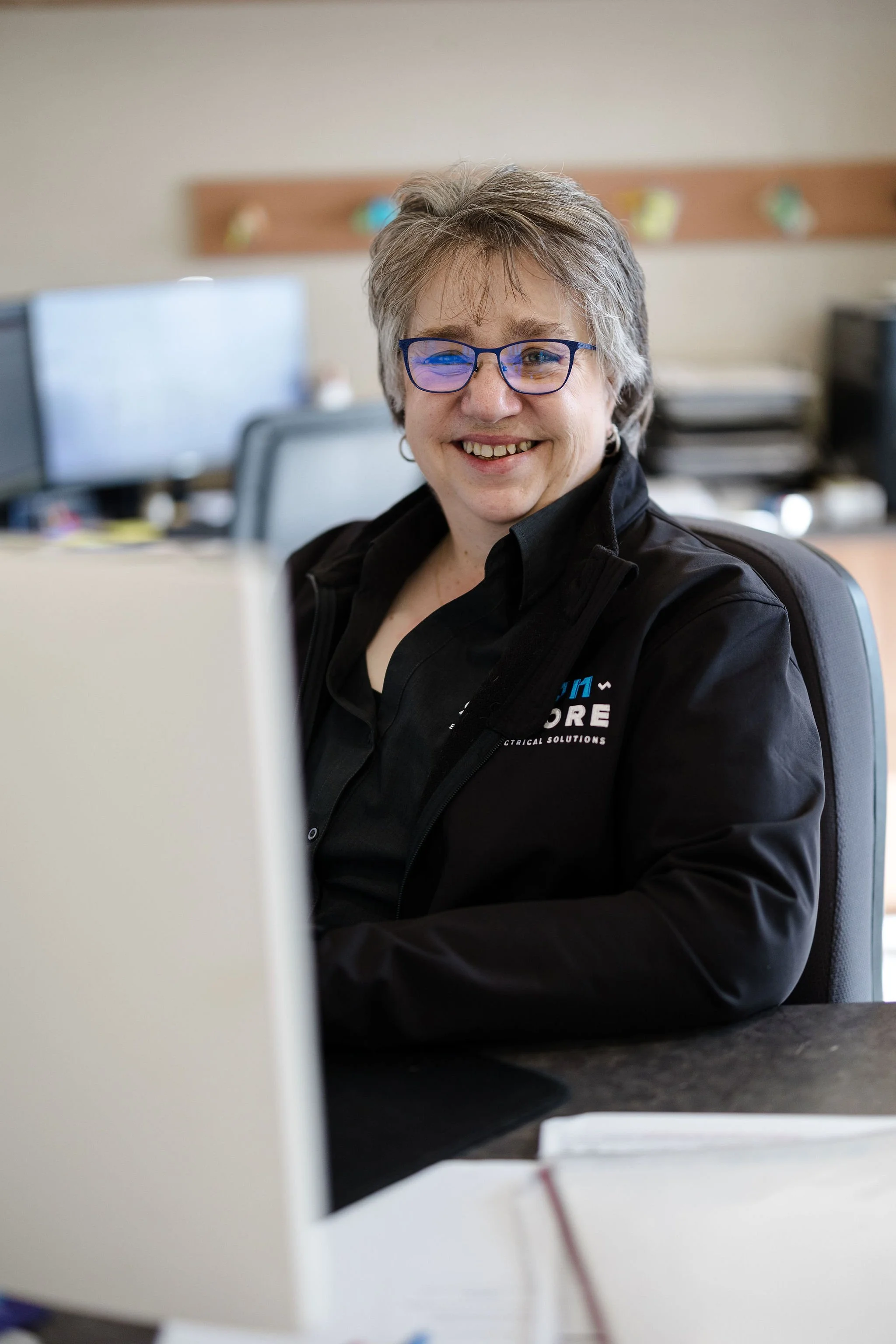 Smiling woman with gray hair and blue glasses sitting at a desk in an office