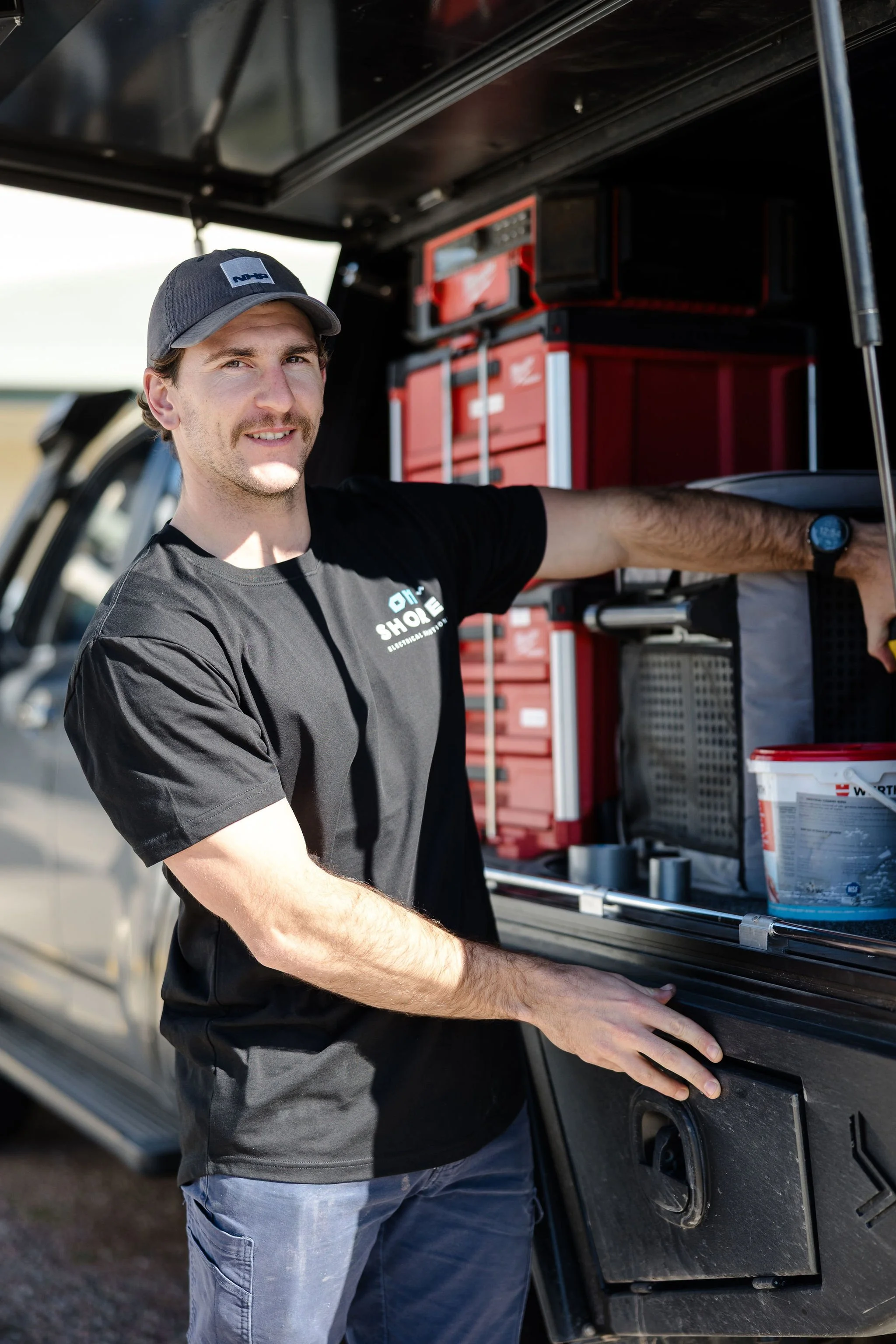 Man wearing a black cap and shirt stands beside an open utility truck filled with red toolboxes and equipment.