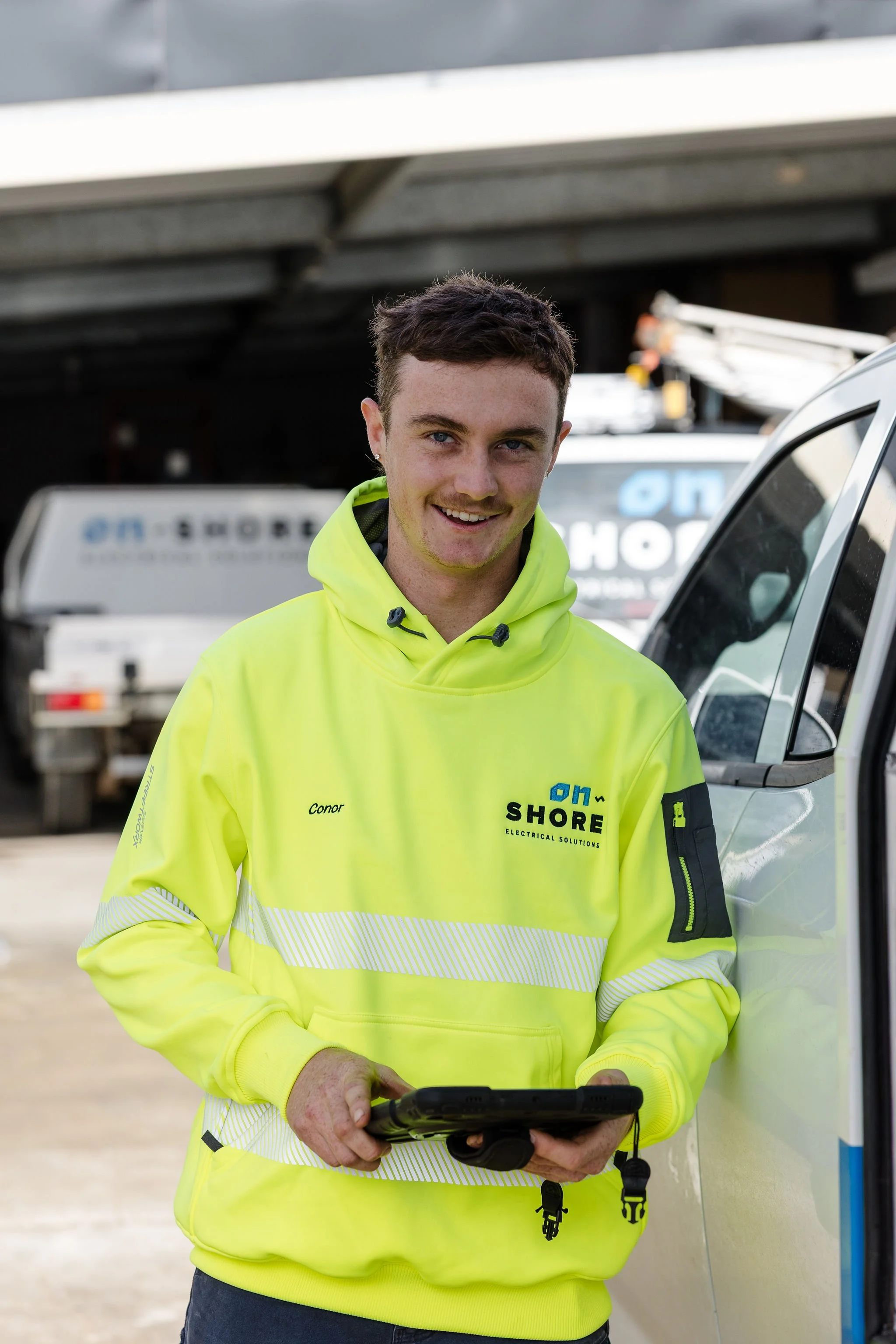 Smiling electrical worker in a high-visibility yellow hoodie holding a tablet beside a service vehicle