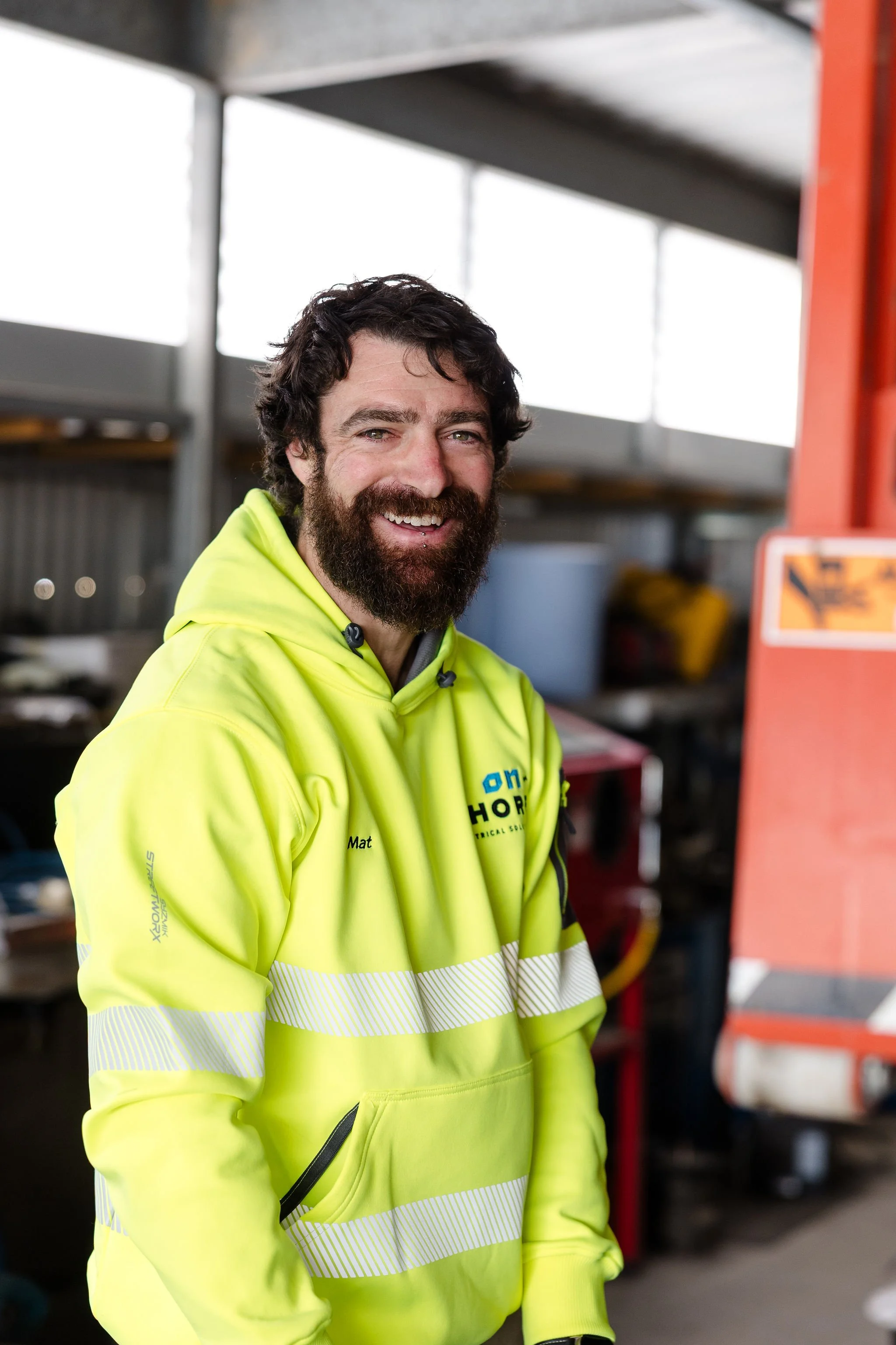 Smiling bearded man in a high-visibility neon yellow work hoodie inside an industrial workshop