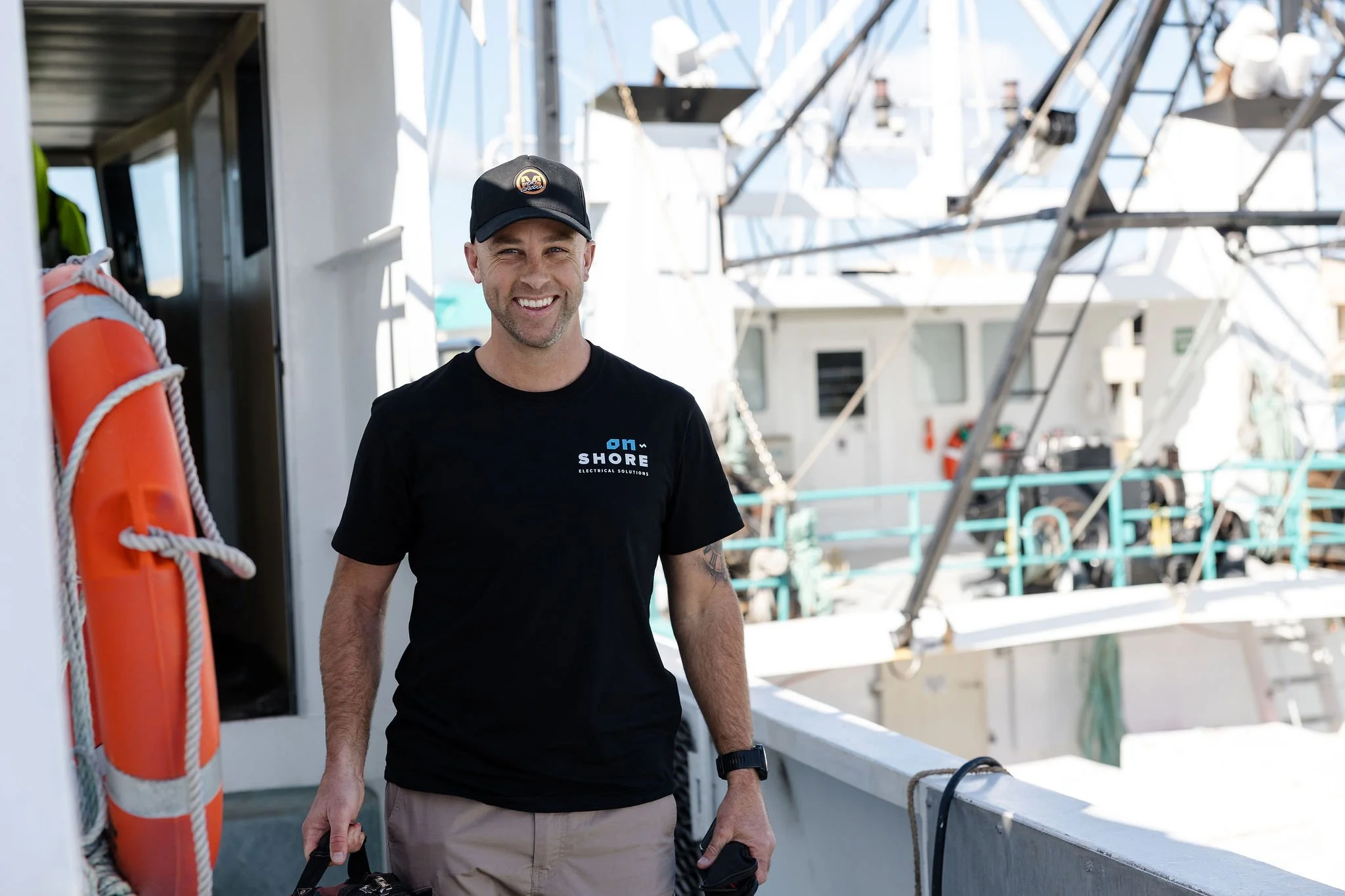 Smiling man in a black cap and branded shirt standing aboard a boat beside an orange life ring