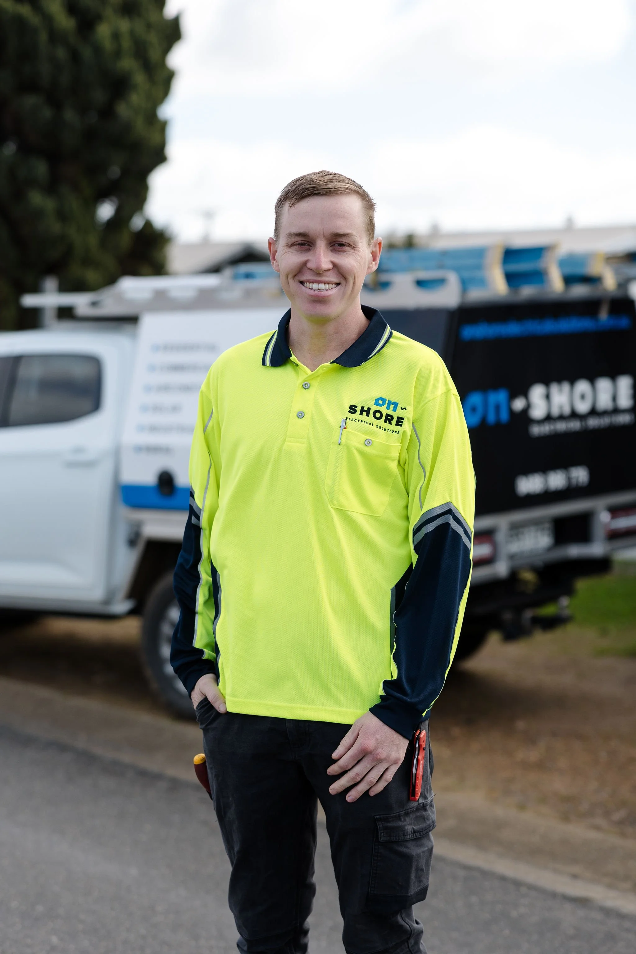 Smiling electrical technician in a high-visibility Shore Electrical Solutions uniform standing in front of service vehicles.