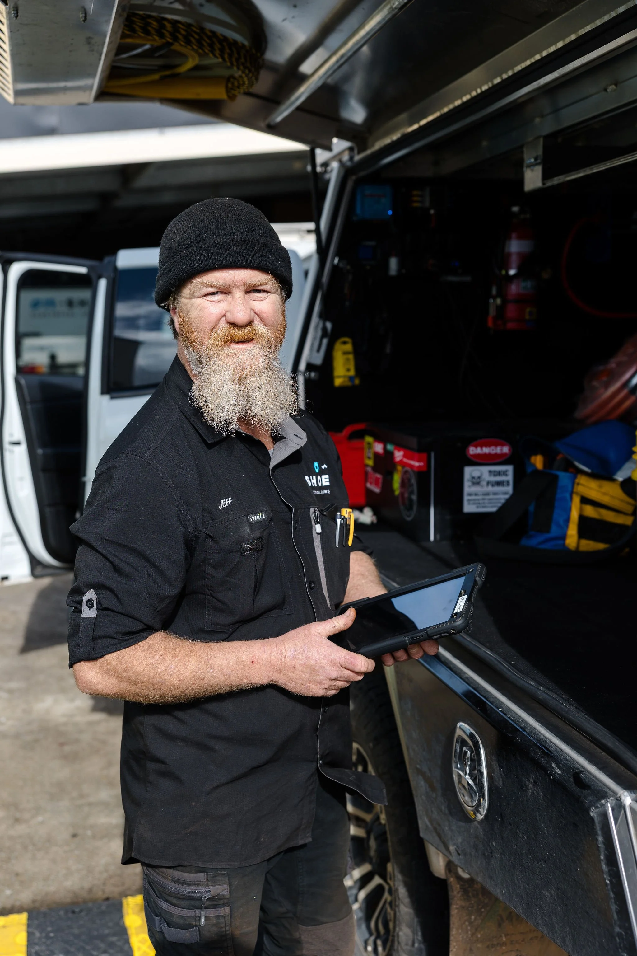 Bearded automotive technician in a black beanie holding a tablet beside an open vehicle service truck.