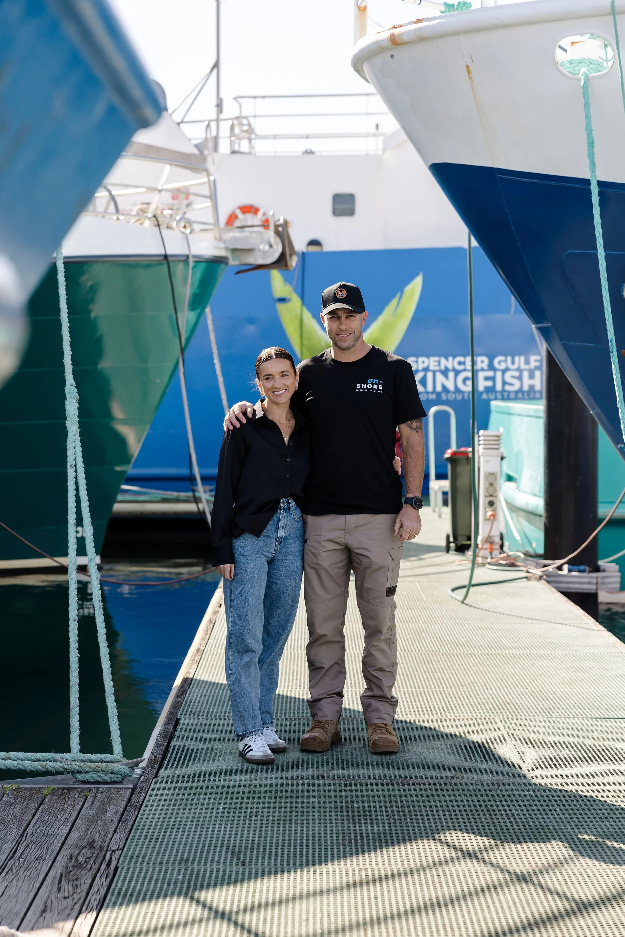 Two people pose on a marina dock between fishing boats, with a fishing tournament banner in the background.
