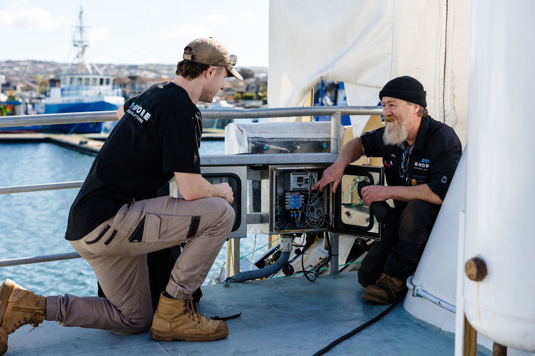 Two electricians work on an open electrical control panel aboard a boat in a harbor.