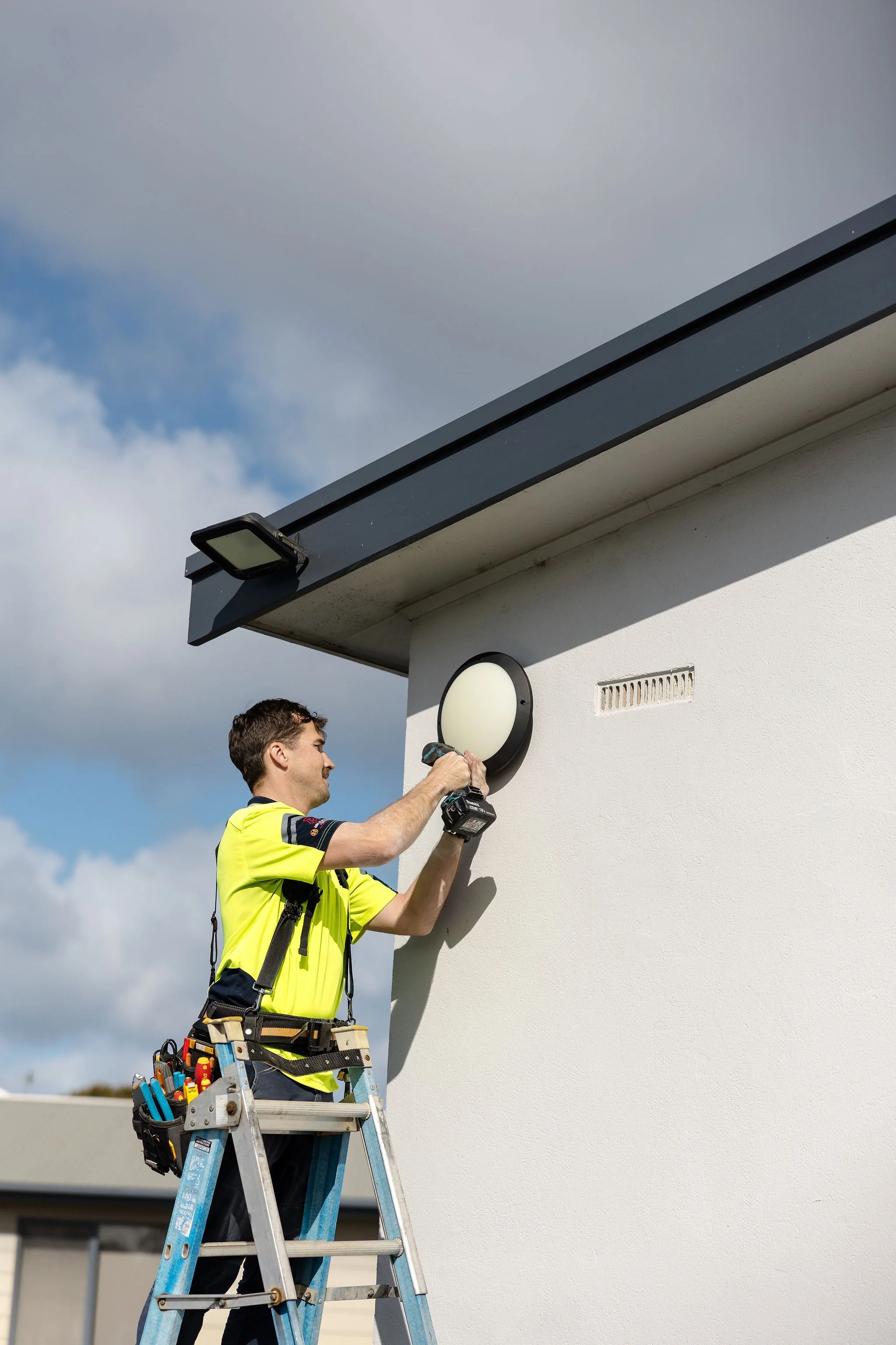 Electrician on a ladder installing an exterior wall light beneath a building eave