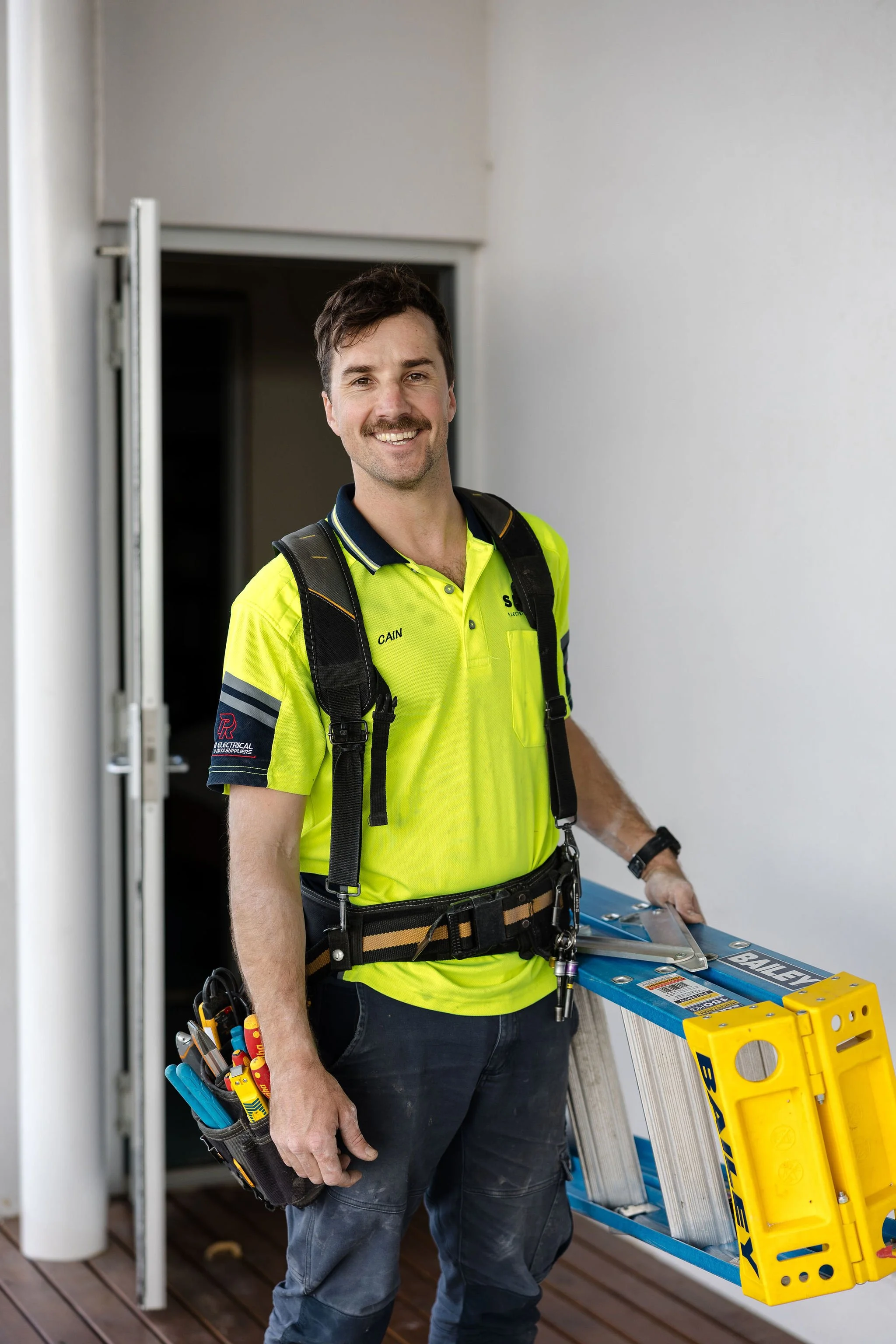 Smiling electrician in a high-visibility shirt and safety harness standing beside an extension ladder at a doorway