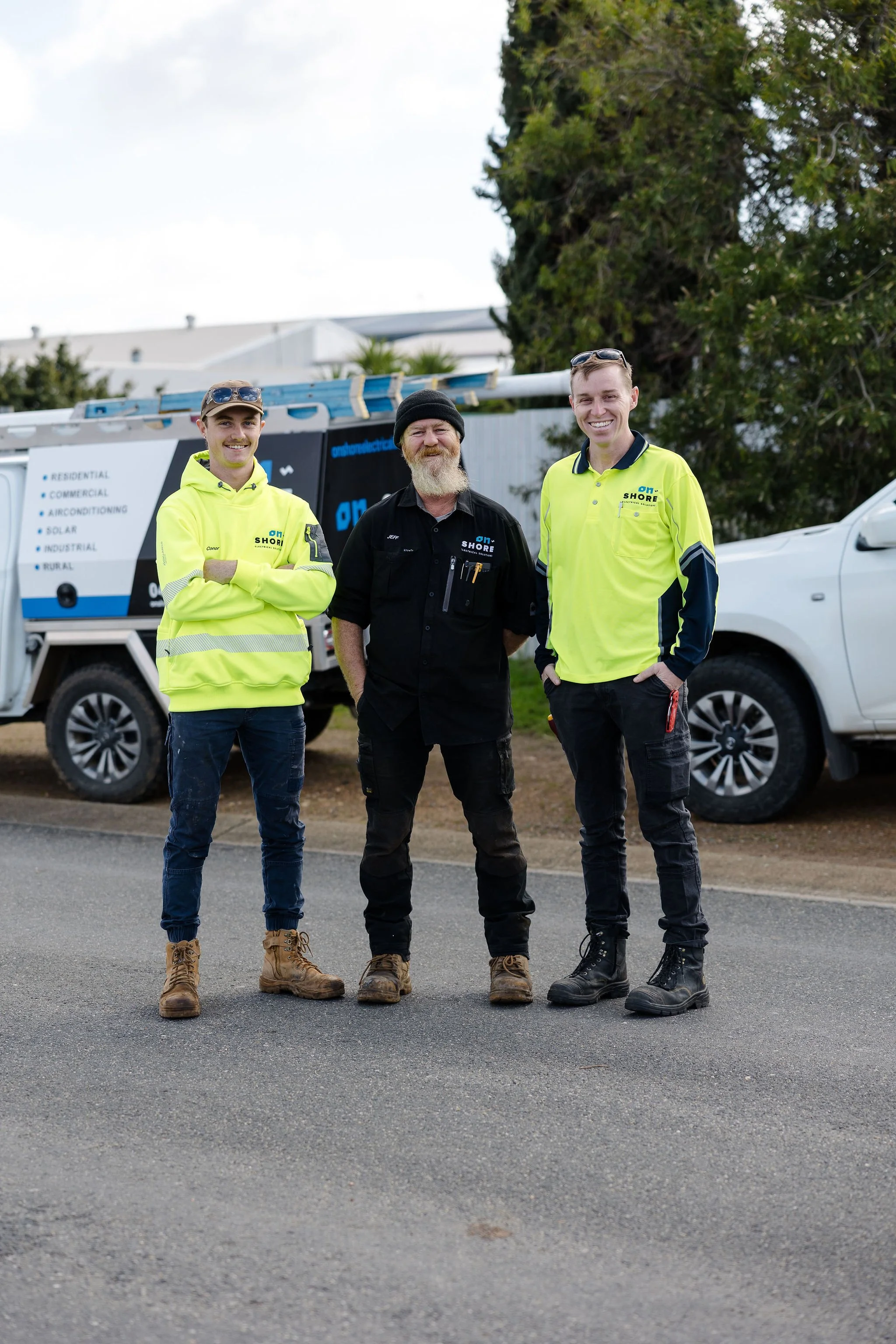 Three electrical workers pose in front of a service truck, wearing high-visibility work shirts and boots.