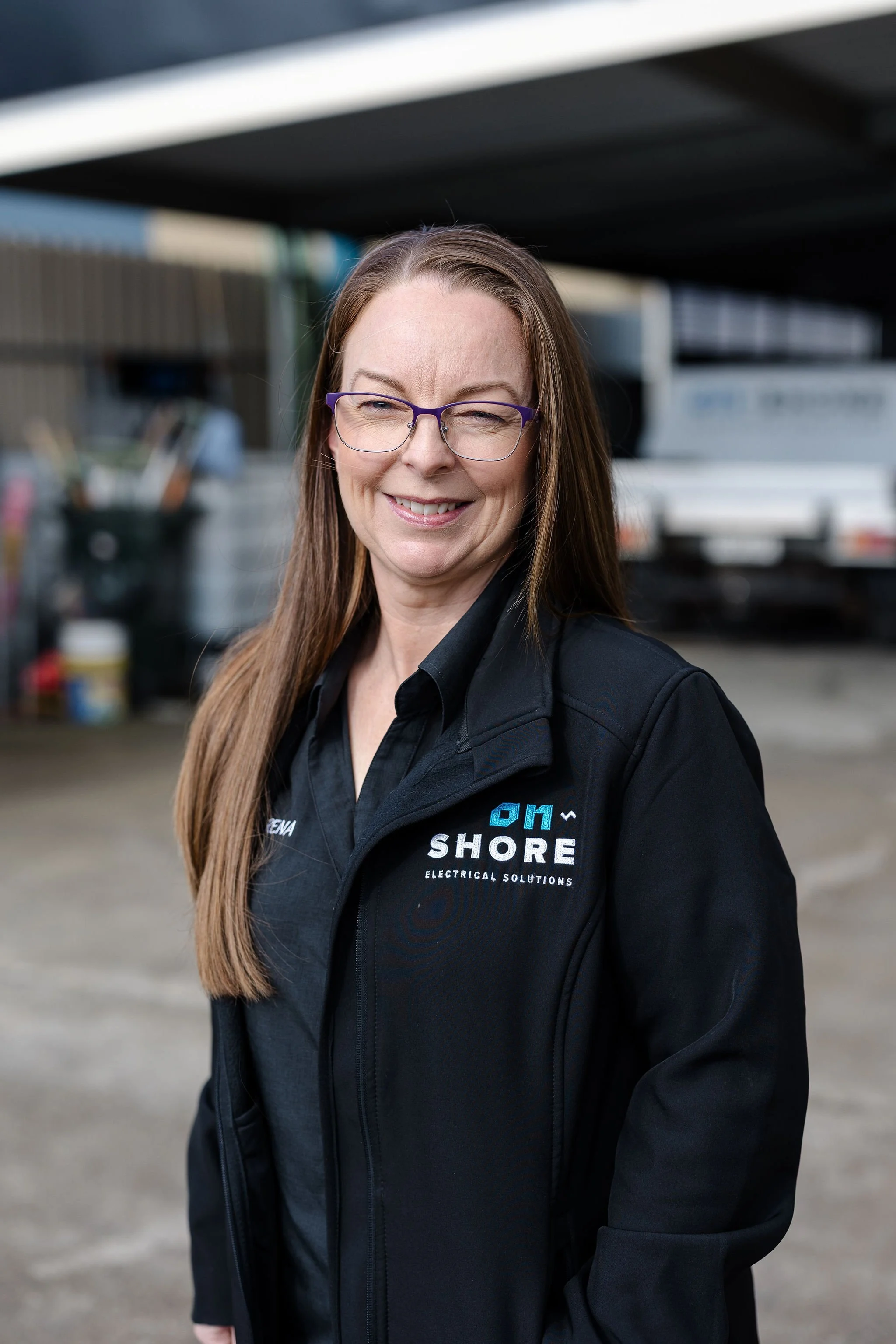 Smiling woman wearing purple glasses and a black Shore Electrical Solutions jacket in a workshop