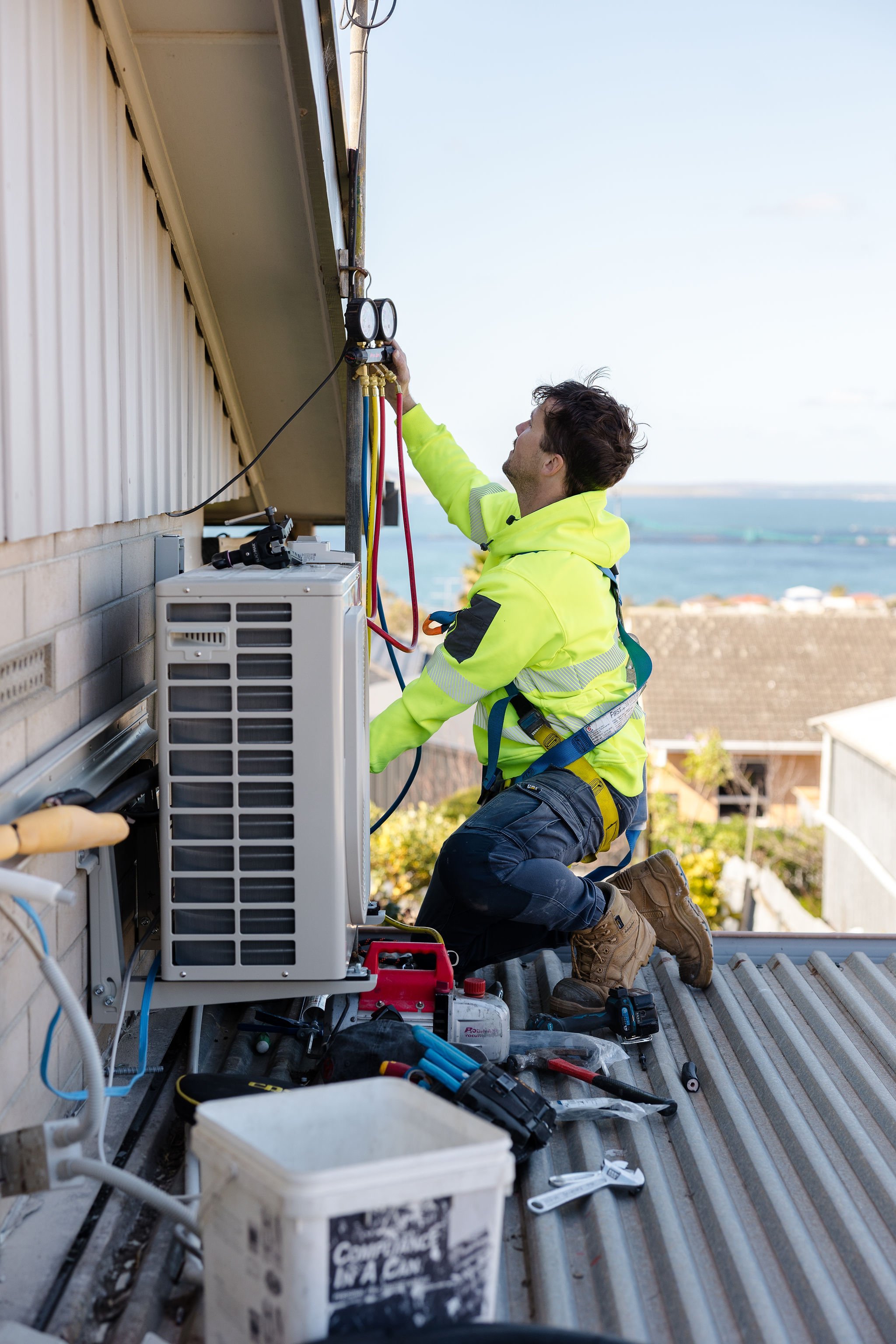 HVAC technician in a safety harness servicing a rooftop air-conditioning unit with pressure gauges and hoses