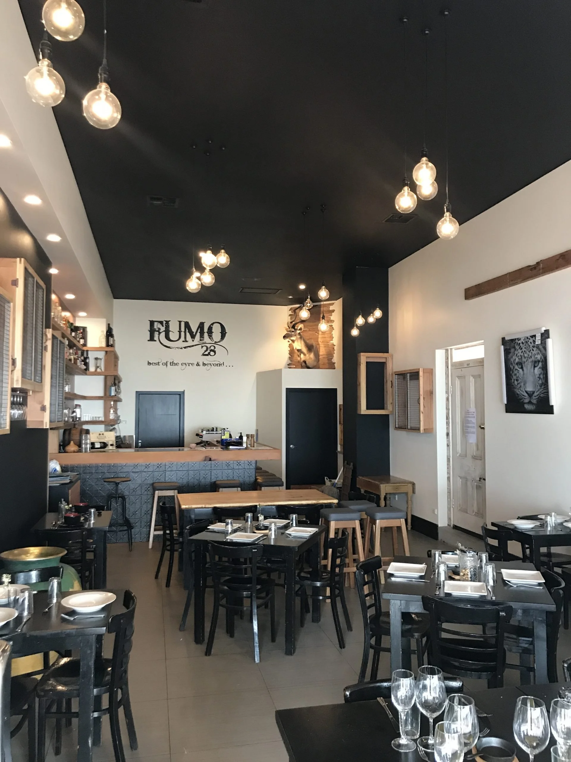Interior of a modern restaurant with black tables, place settings, a bar, pendant lights, and rustic wood-and-animal decor.