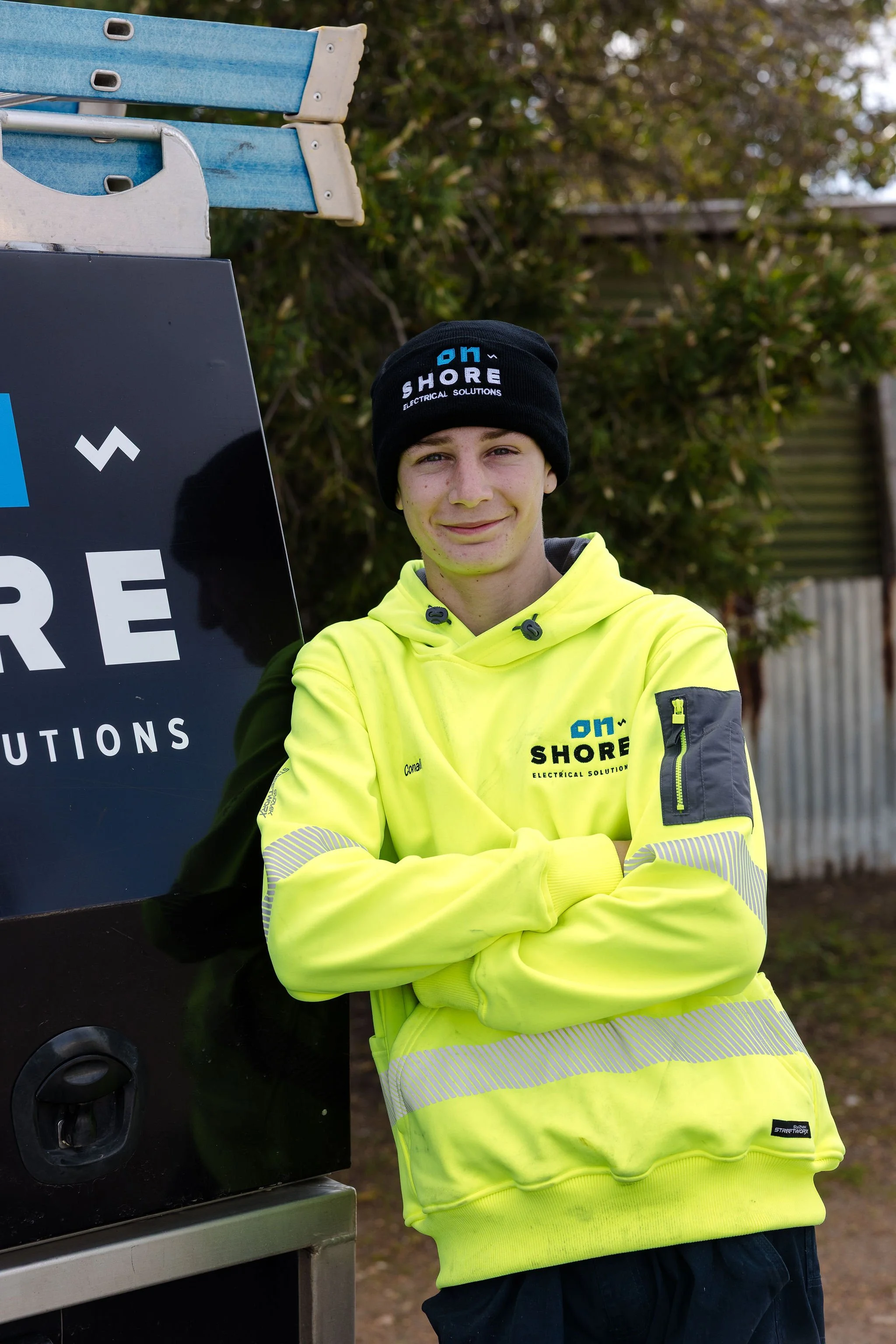 Electrician wearing a high-visibility hoodie and branded beanie beside a work trailer outdoors