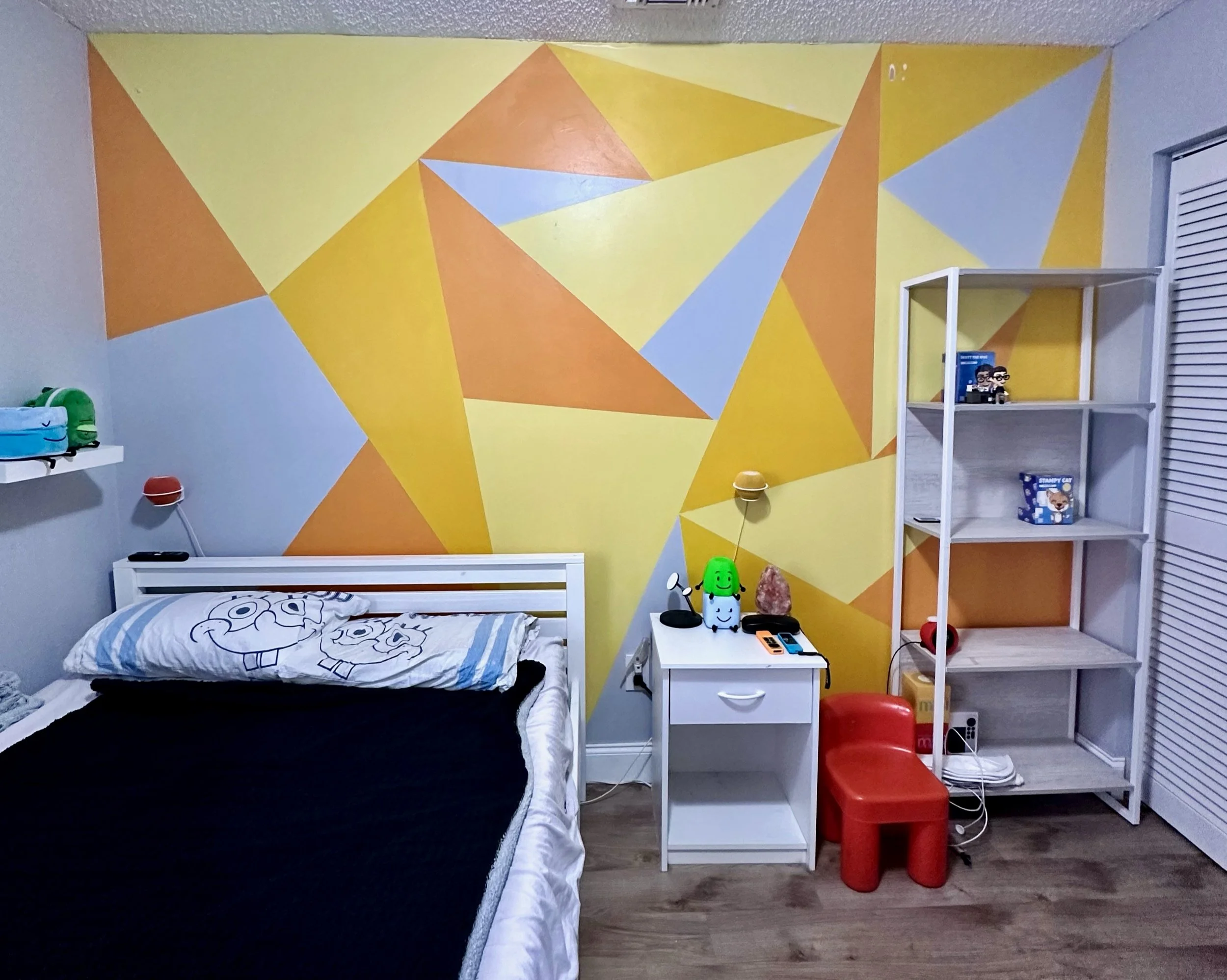 Bedroom with a geometric yellow, gray and orange wall mural, white bed with a cartoon character pillow, a white nightstand with toys and a salt lamp, and a white bookshelf with various items. Red and yellow chairs on a wood floor.