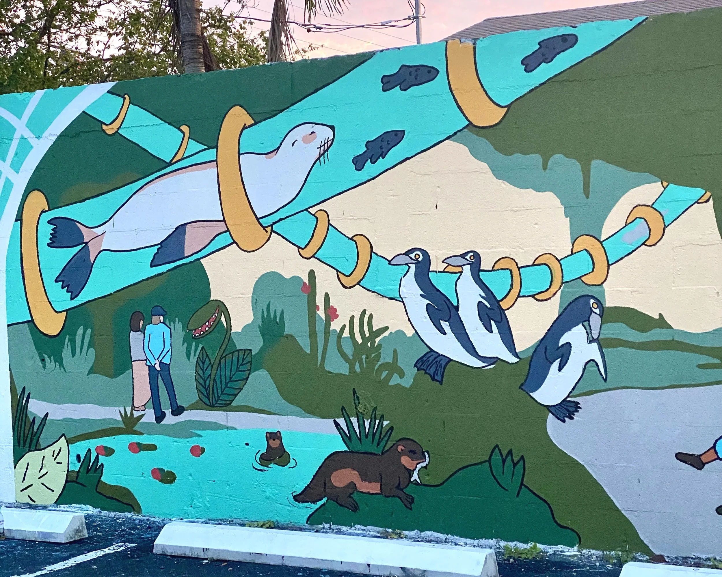 Colorful mural featuring a large fish in a tube with rings, four penguins hanging under the tube, two humans, and landscape elements like trees and hills.