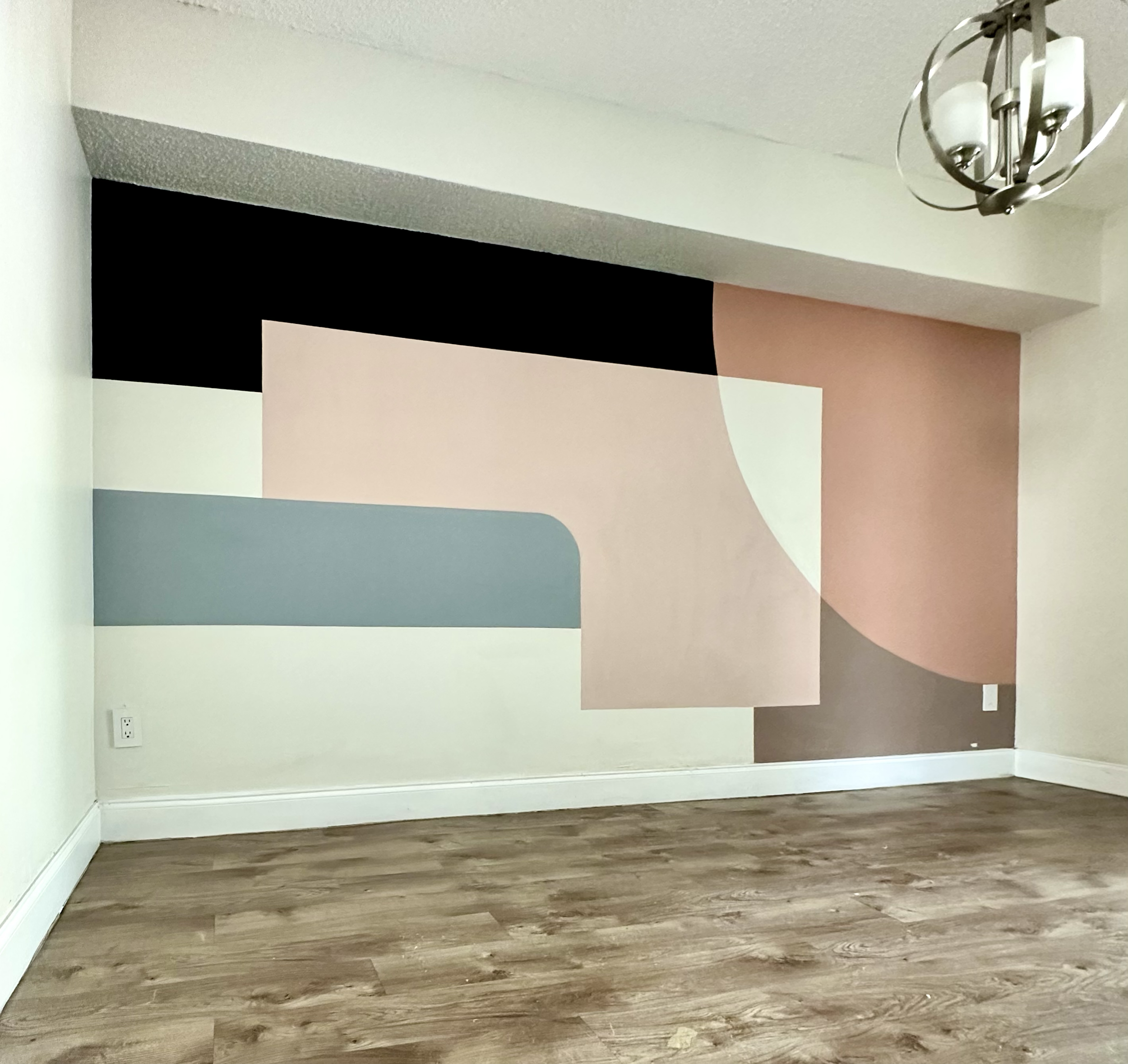 A room with a wooden floor and a wall painted with abstract geometric shapes in black, pink, beige, white, and blue. There is a modern light fixture hanging from the ceiling.
