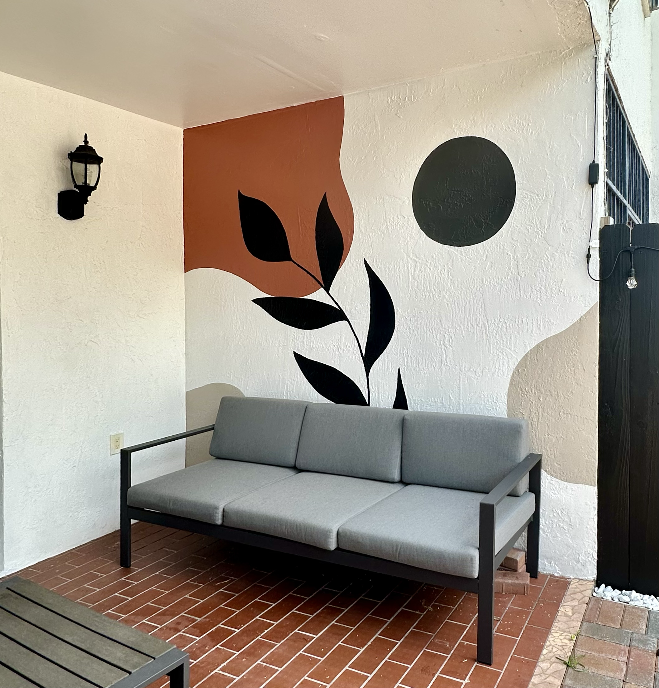 Outdoor seating area with a grey outdoor couch in front of a white wall decorated with abstract black, brown, and beige shapes with a vine and a black circle, and a black lamp on the wall.